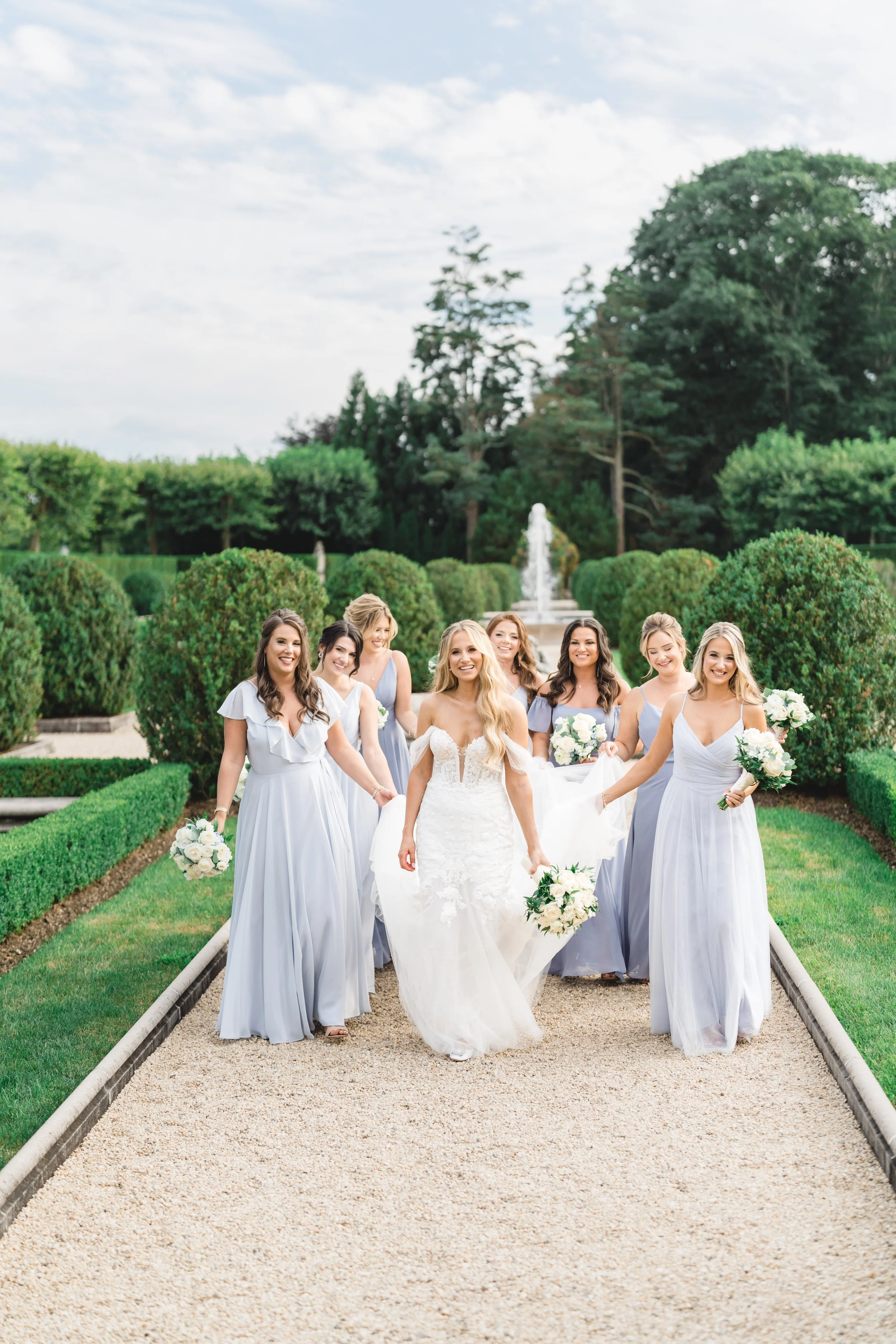 Bride walking with bridesmaids in soft blue dresses along garden path at Oheka Castle
