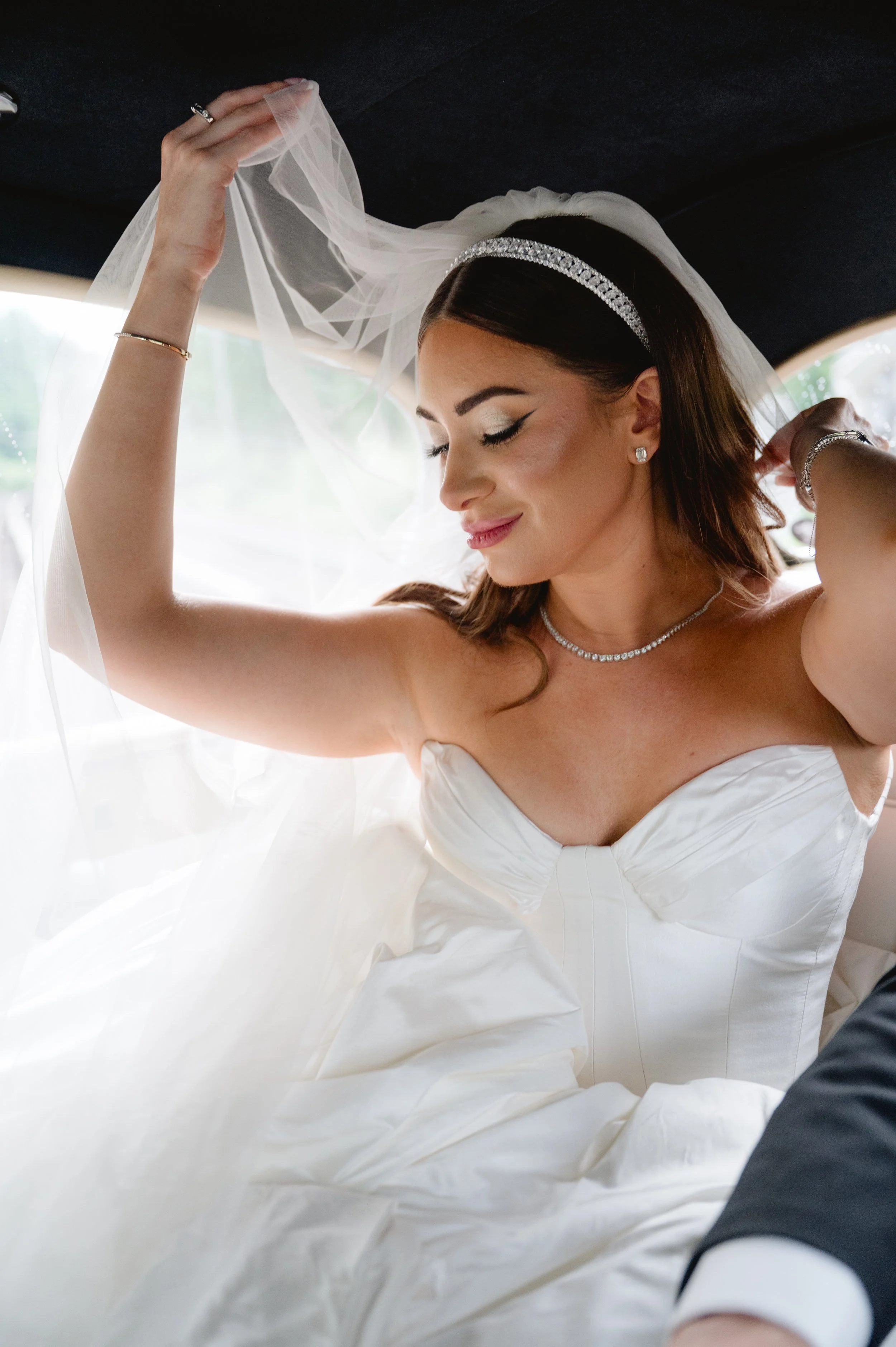 A bride in a white wedding dress and veil, with jewelry, smiling with eyes closed, sitting inside a vehicle.