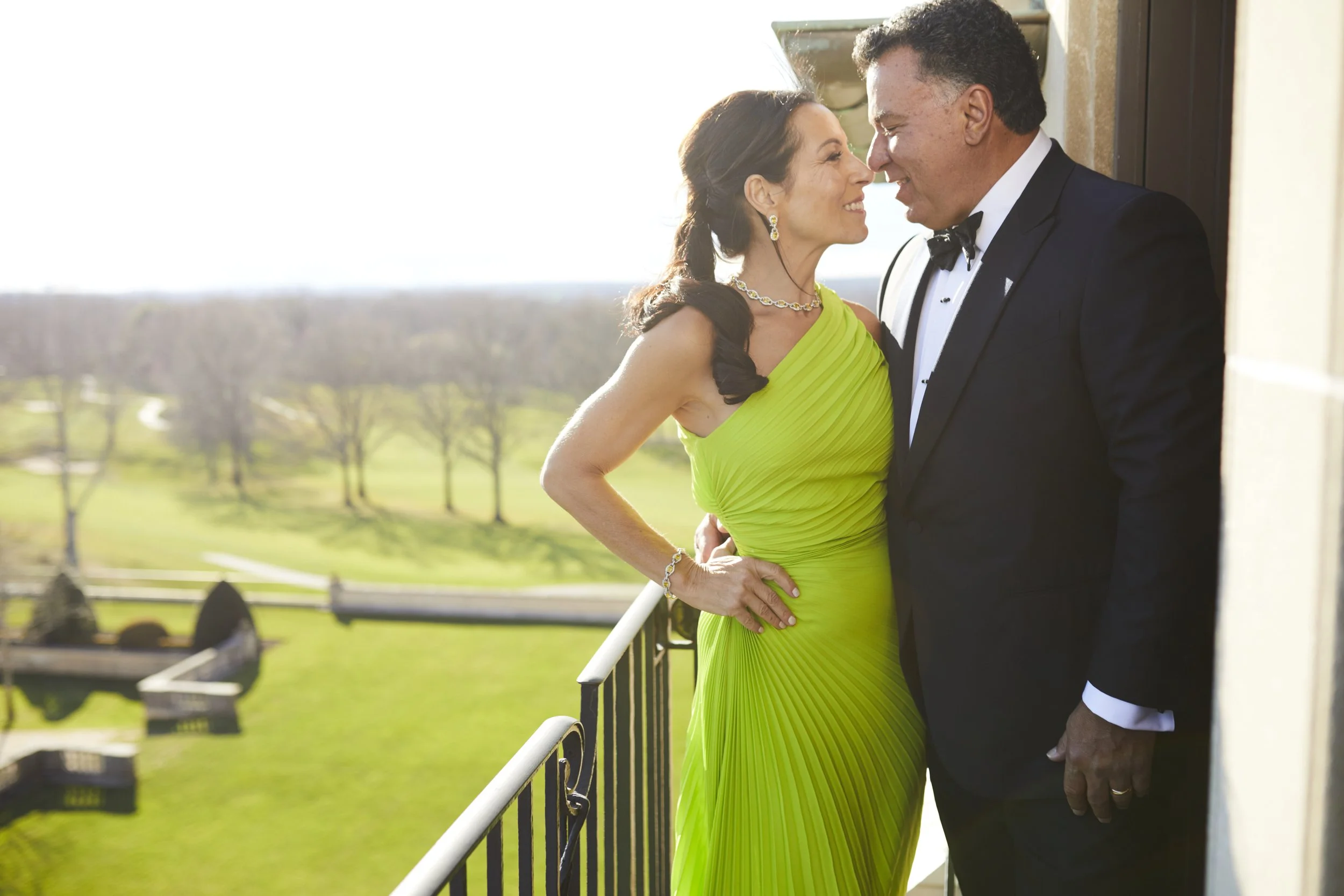 A couple dressed in formal attire standing close together on a balcony overlooking a green landscape, smiling with their foreheads touching.