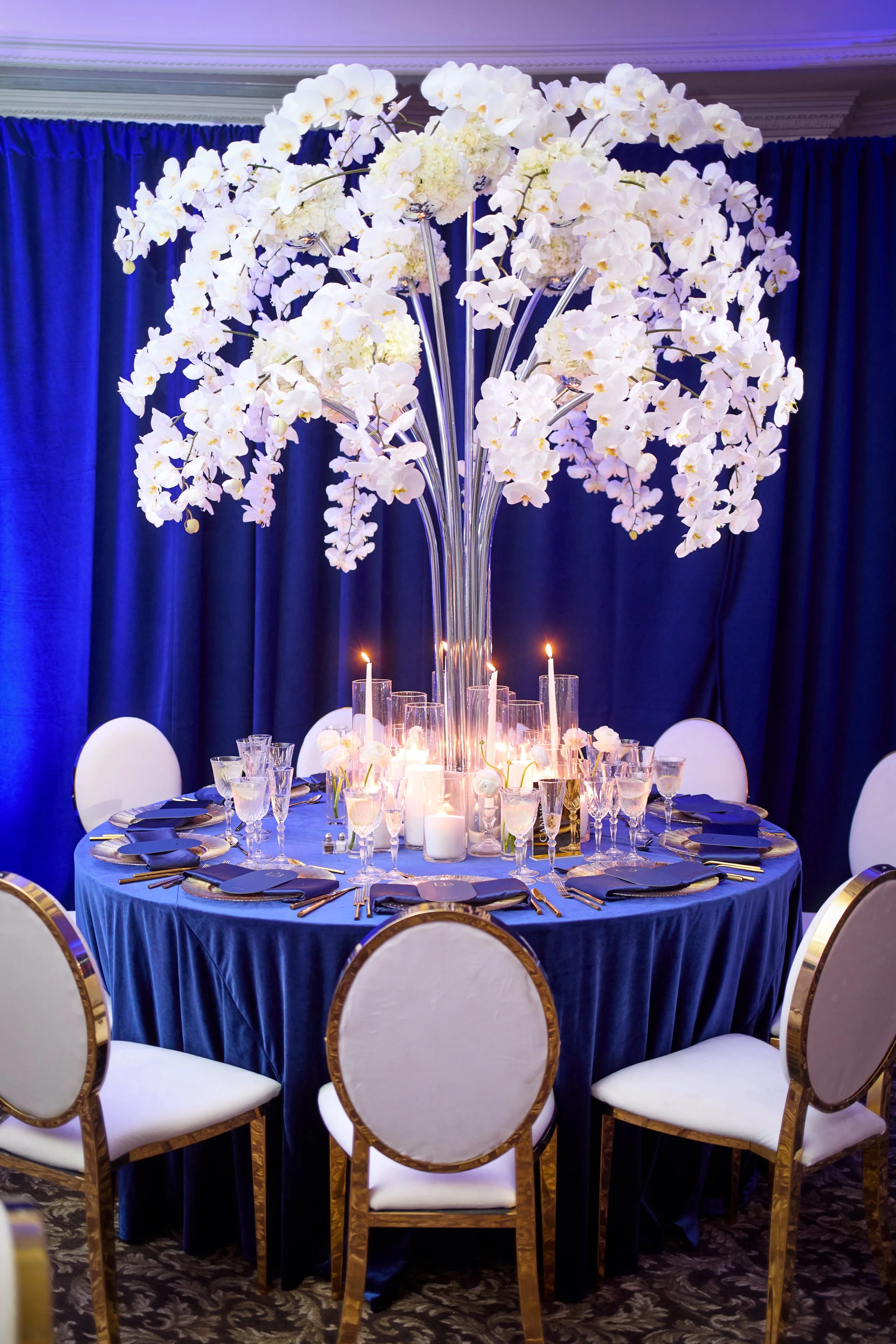 Custom orchid “trees” rose from each table, composed of cascading white phalaenopsis arranged in a sculptural, branching form. Set against deep blue velvet and gold accents, the design felt both architectural and organic—bringing height, movement, an