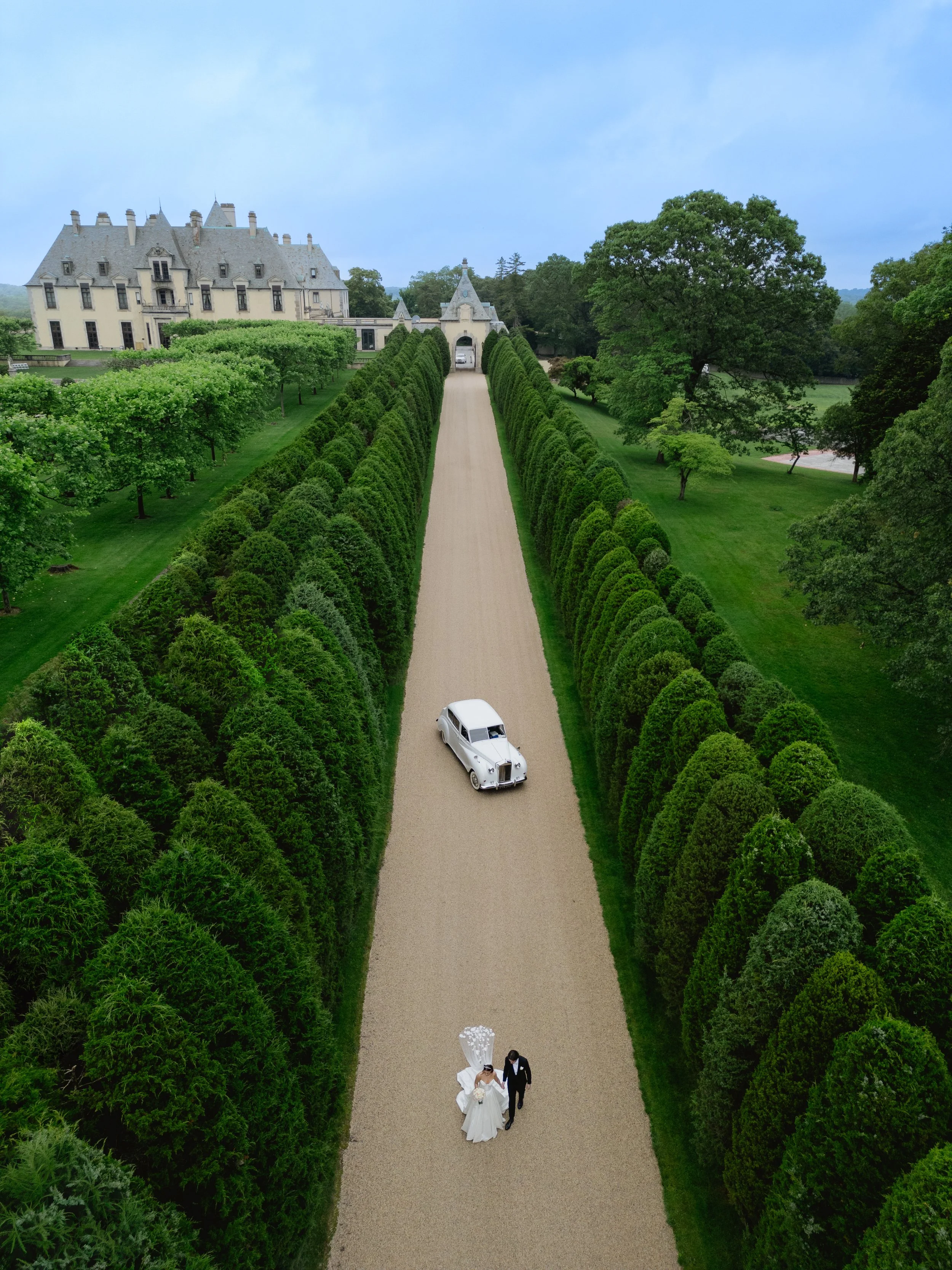 A bride and groom walking down a gravel path at a wedding venue, with a vintage car nearby and a large castle-like building in the background, surrounded by green trees and well-manicured bushes.