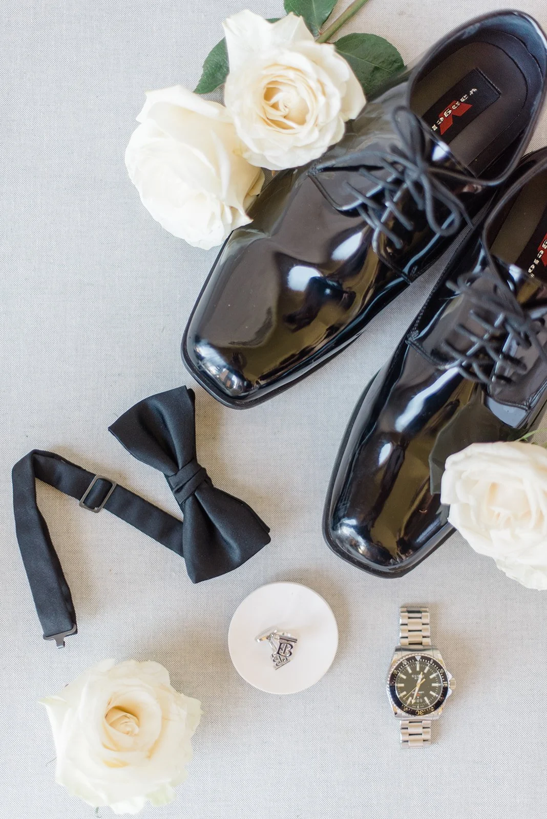 Black shiny oxford shoes, white roses, black bow tie, wristwatch, small silver jewelry on a white surface.