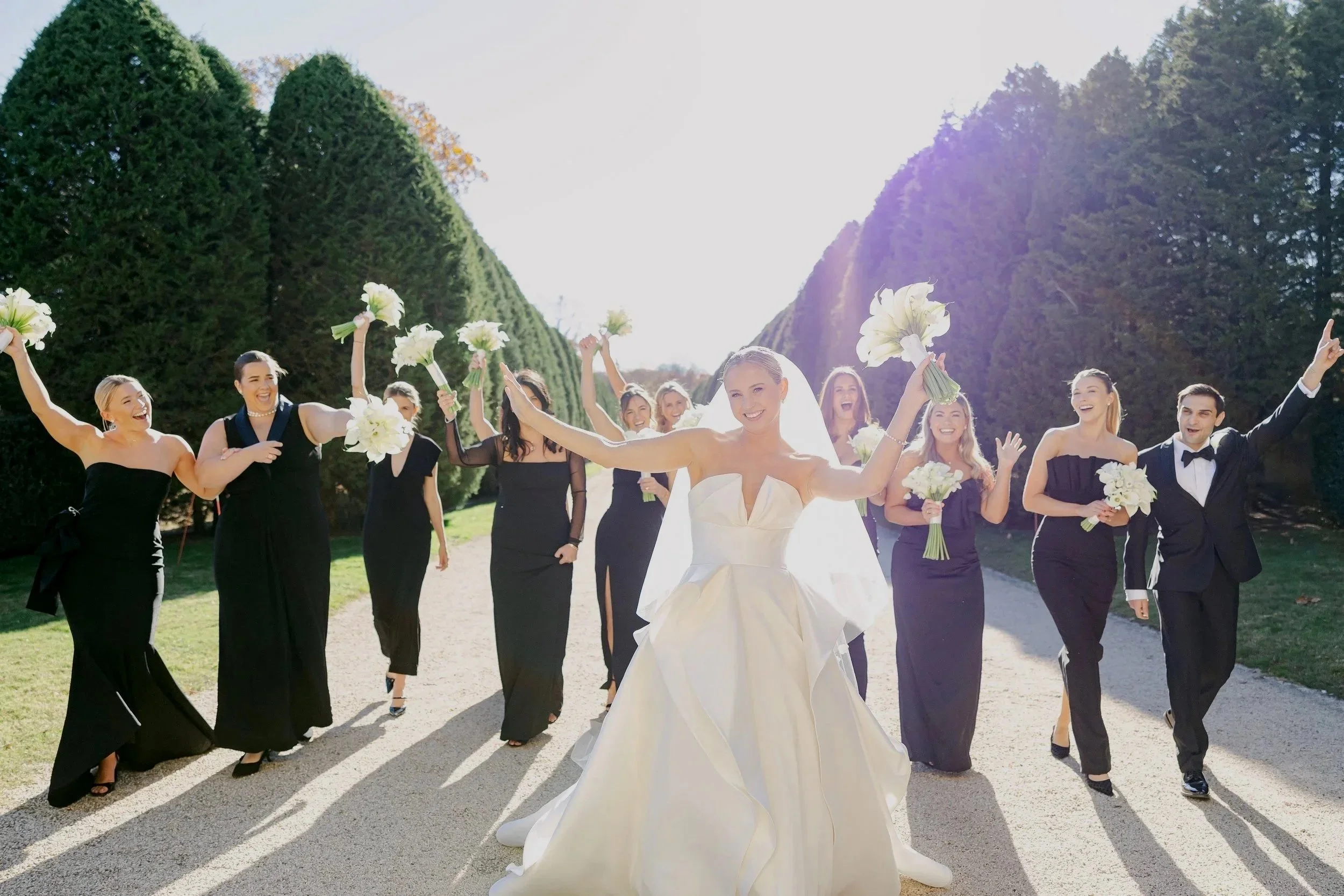 Bride in white wedding gown and veil smiling with bridesmaids and groomsmen holding bouquets outdoors on a sunny day, walking on a gravel path between tall green trees.