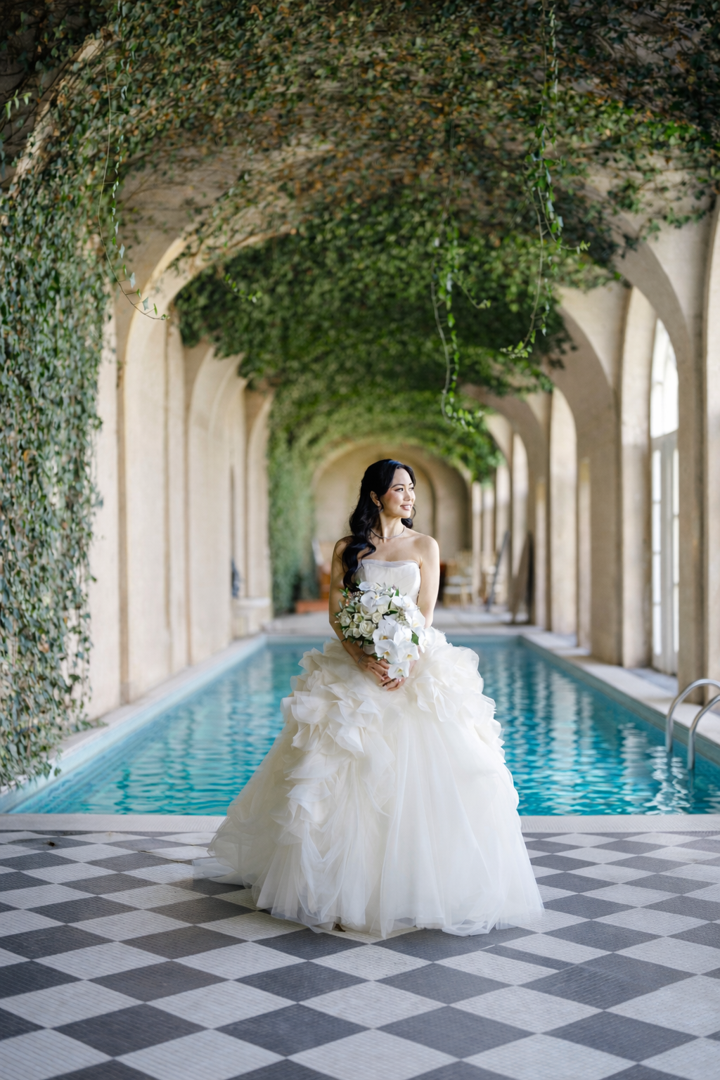 Bride in a white wedding gown holding a bouquet of white flowers by an indoor swimming pool with arched windows and vine-covered ceiling.