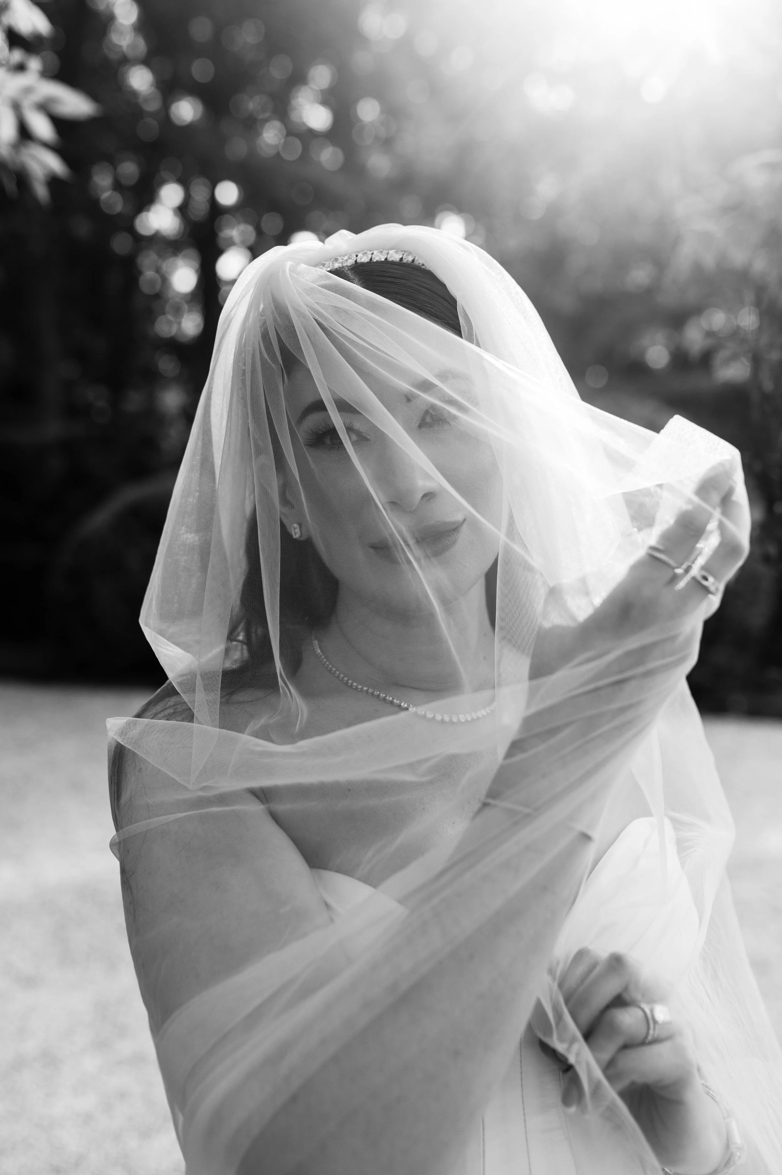 A bride in a wedding dress and veil outdoors, smiling behind the veil, with sunlight and trees in the background.