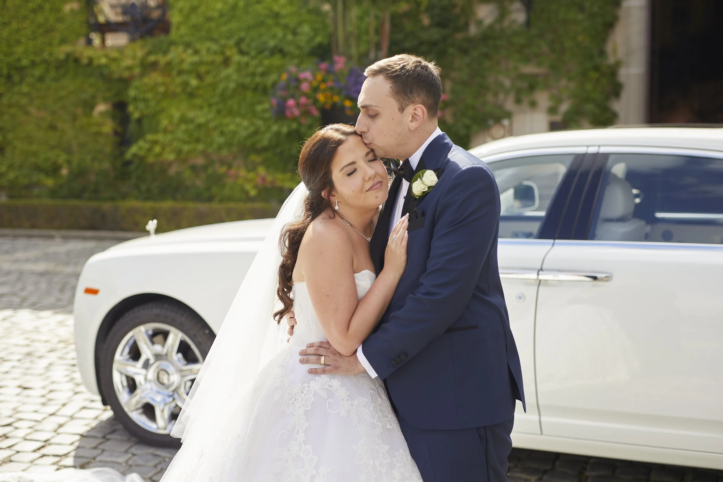 Anthony kissing Samantha on the forehead beside a vintage white Rolls-Royce outside Oheka Castle during the wedding celebration.