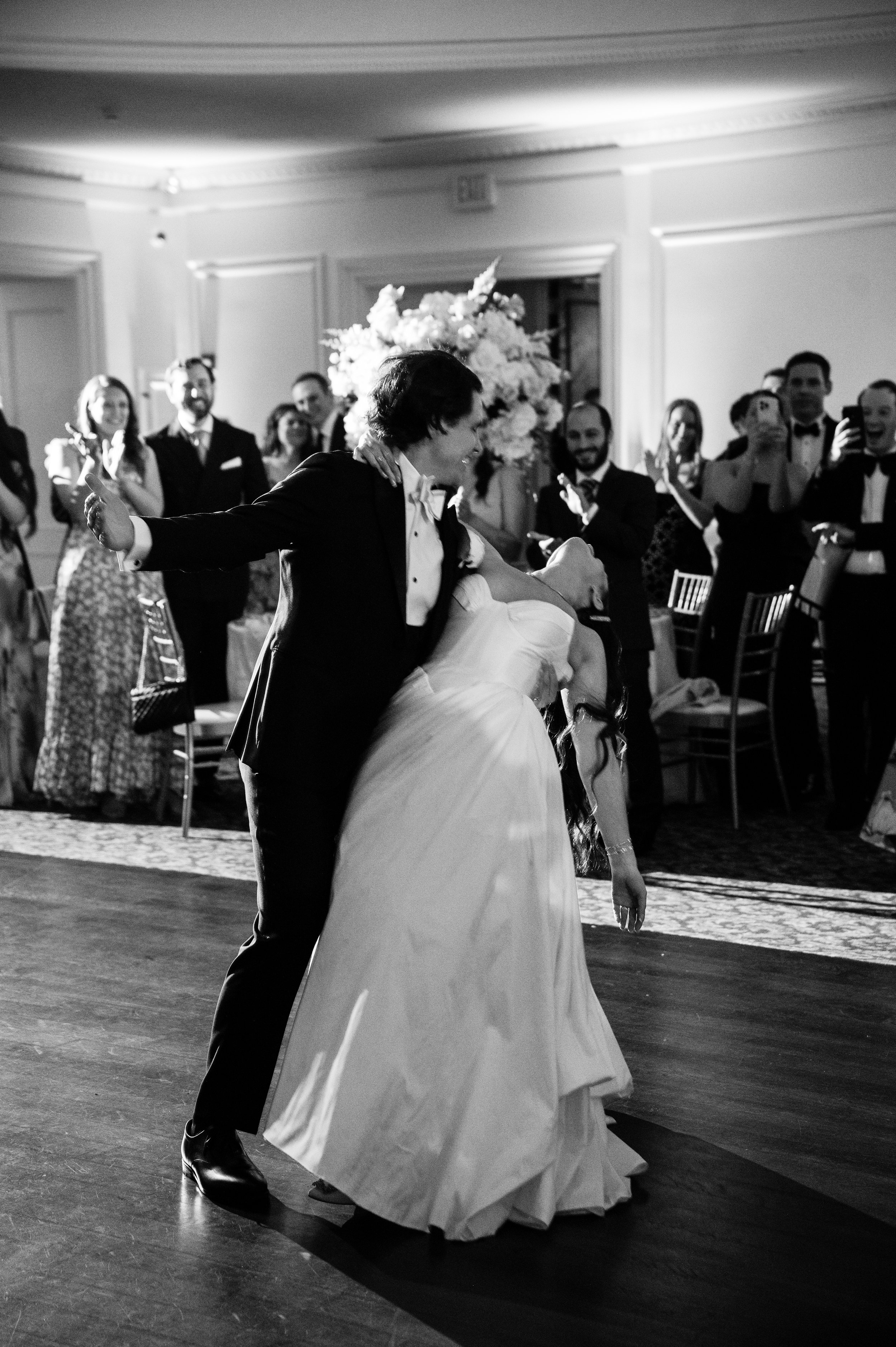 A couple dancing at a wedding reception, with the bride in a white gown and the groom in a tuxedo, surrounded by guests clapping and taking photos.