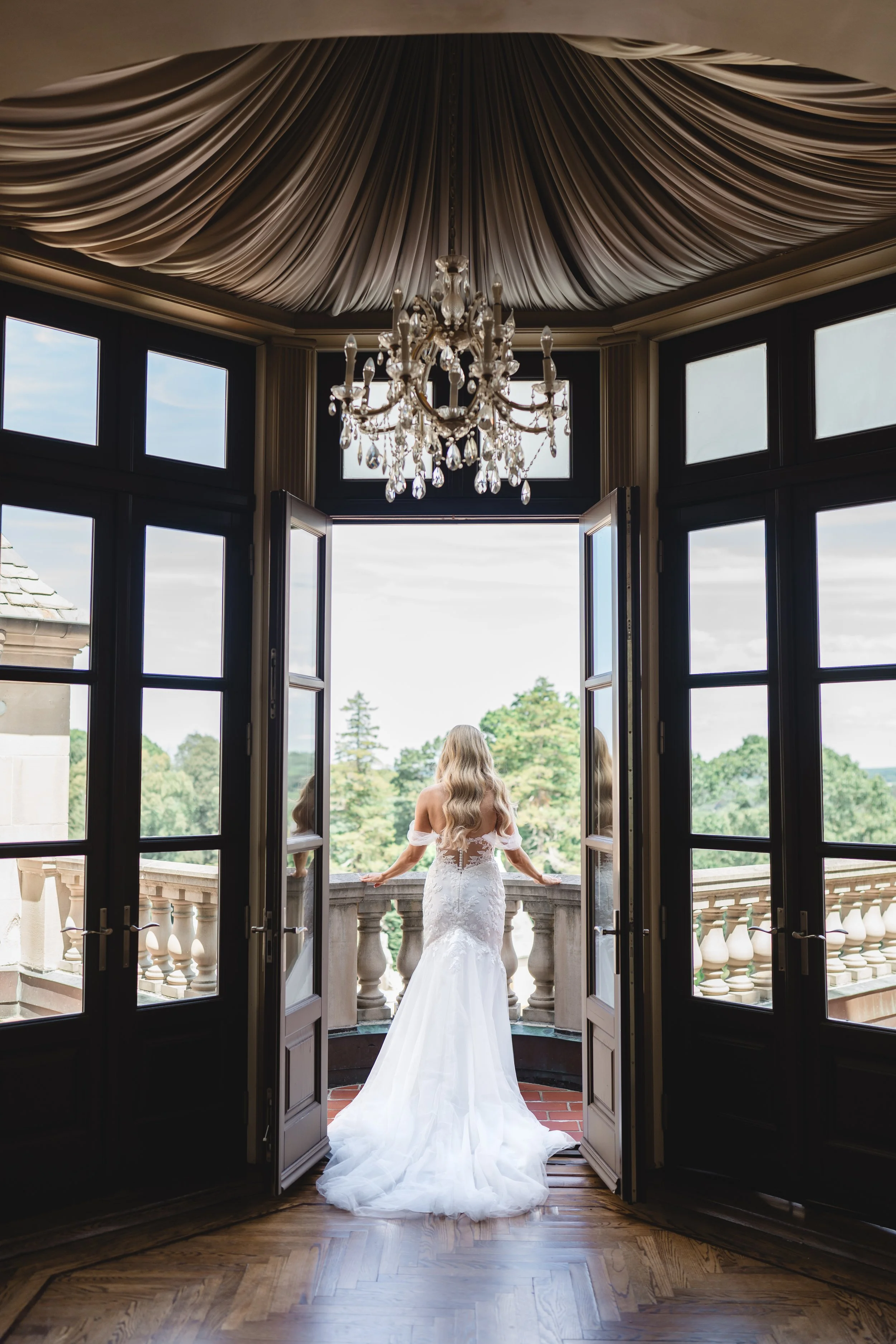Bride standing on balcony at Oheka Castle with open doors and chandelier framing the scene