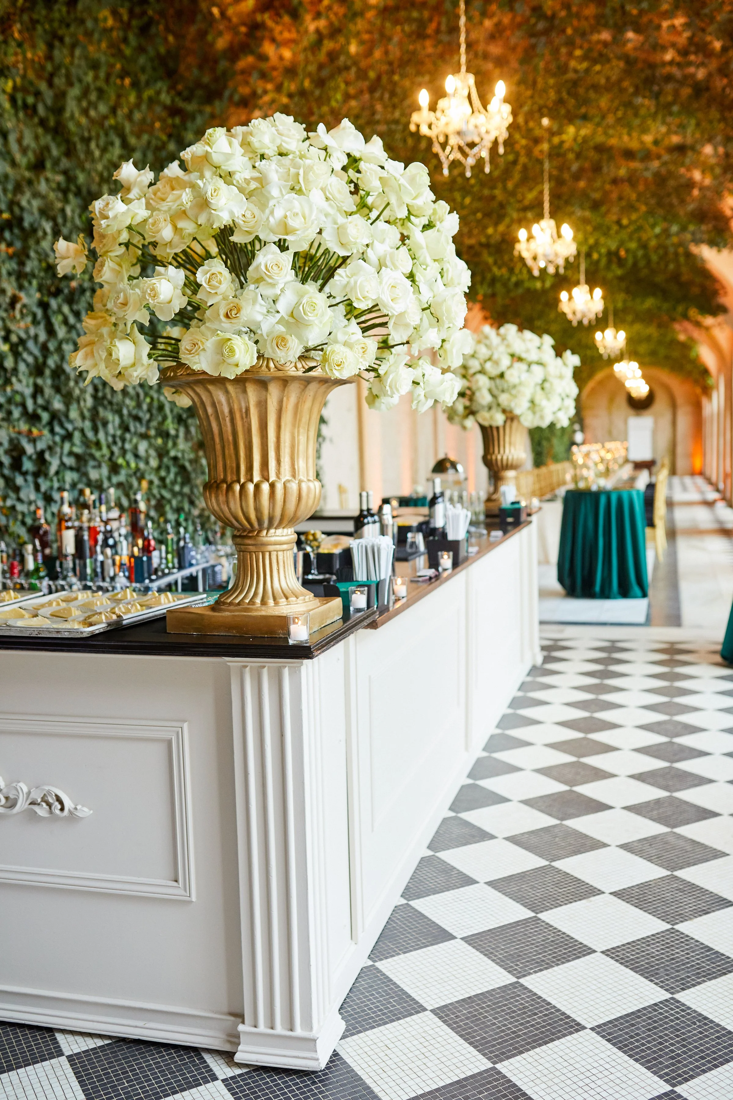Elegant indoor venue with large white floral arrangements in gold vases on a white counter, chandeliers hanging from the ceiling, a wall covered in greenery, black and white checkered floor, and cocktail bar area with bottles and glasses.