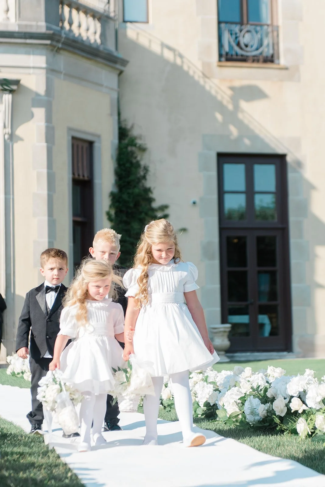 Children dressed in formal attire participating in a wedding ceremony outdoors, with a white aisle runner and floral arrangements, in front of a large building with windows and a balcony.