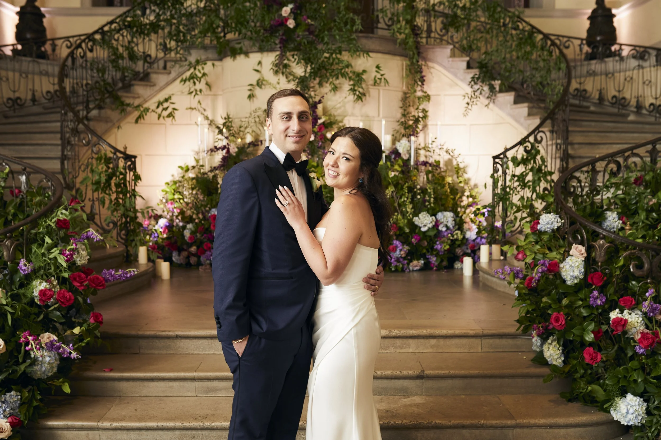 Samantha and Anthony posing on the grand staircase at Oheka Castle surrounded by lush florals, candlelight, and romantic garden-inspired reception décor.