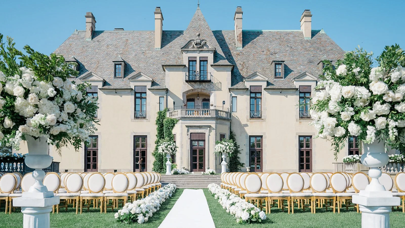 A grand estate prepared for a wedding ceremony, with a white aisle, floral arrangements, and rows of chairs and large flower vases in front of a historic mansion.