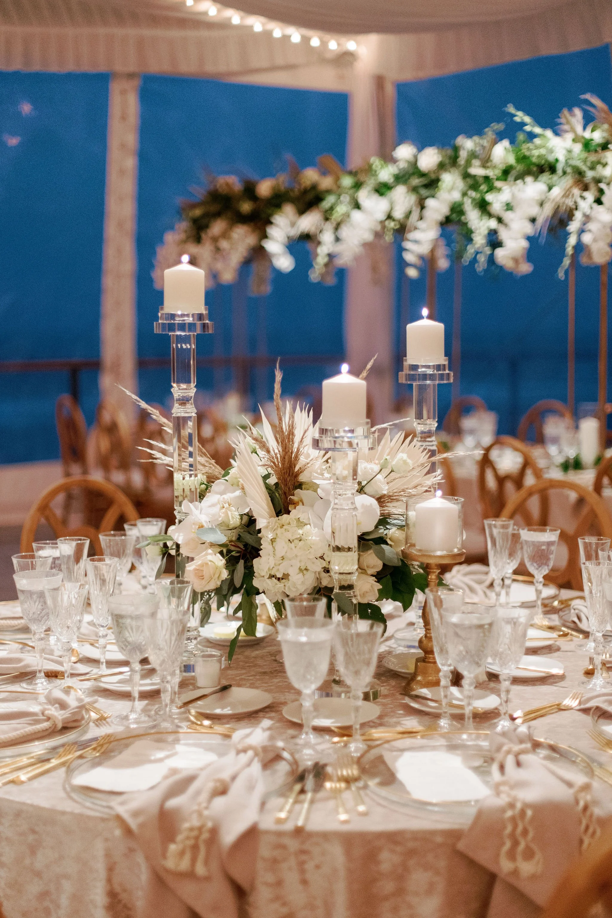 A decorated banquet table at a formal event with white floral centerpieces, candles, and gold accents, set in an elegant venue with blue walls and string lights.