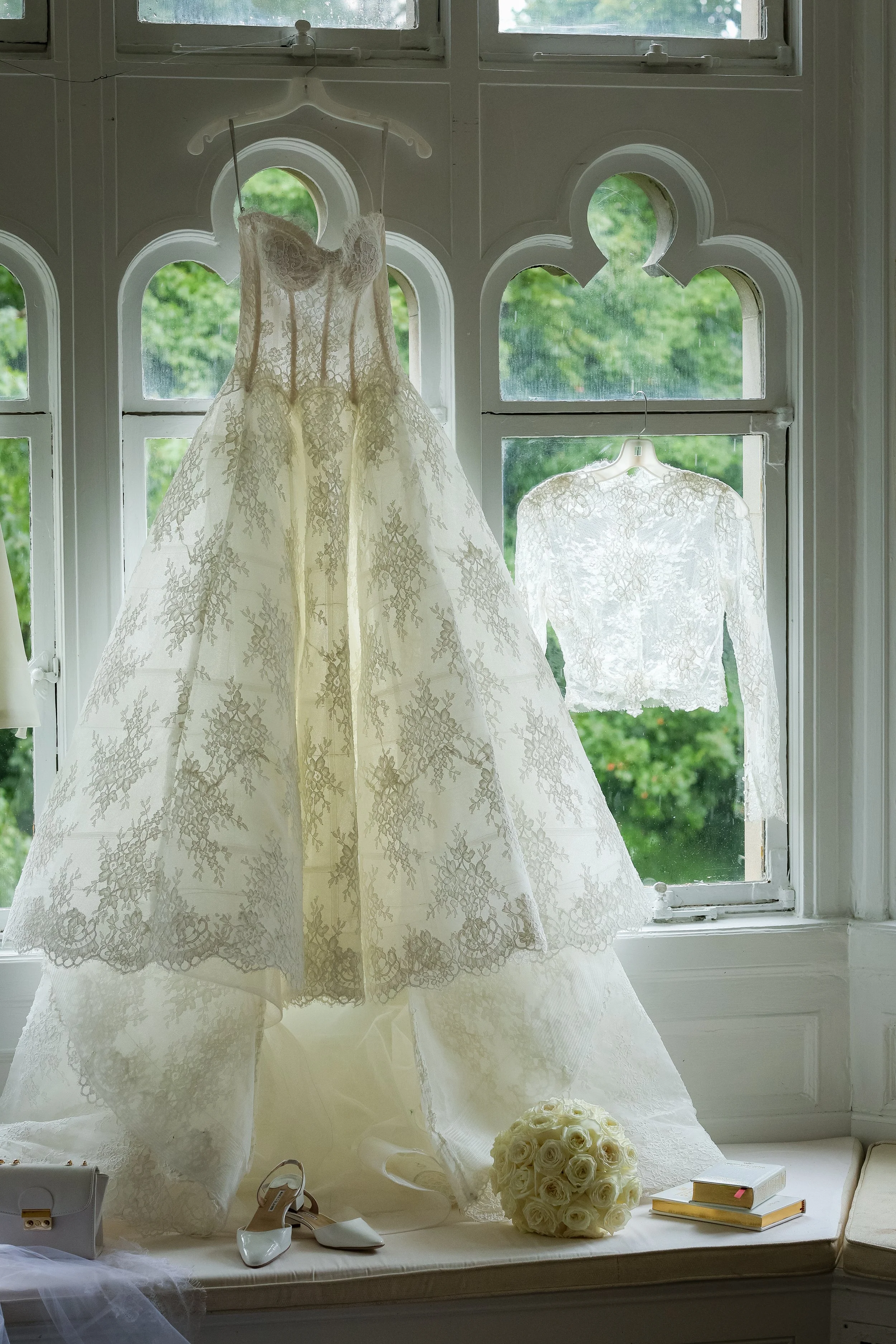 Bridal gown hanging in front of a window, with a bouquet of white roses, a pair of white heels, a clutch, and a few books on the windowsill.