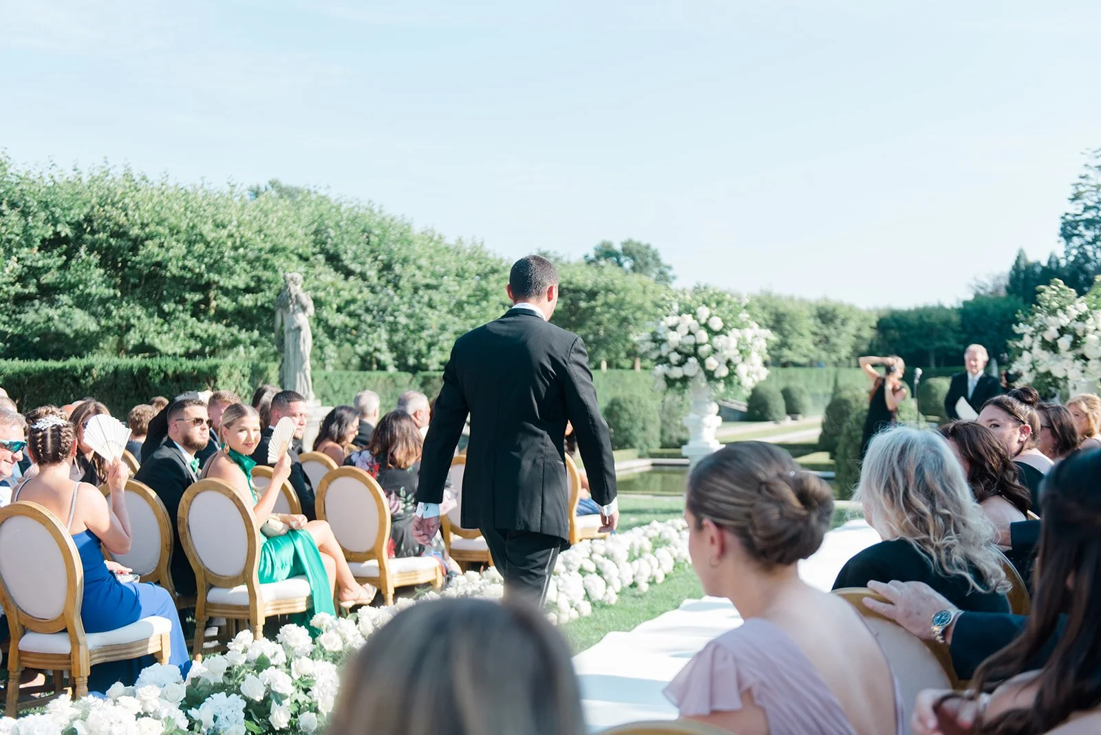 Outdoor wedding ceremony with guests seated, a man in a black tuxedo walking towards the altar, and a garden setting with white flowers and greenery.