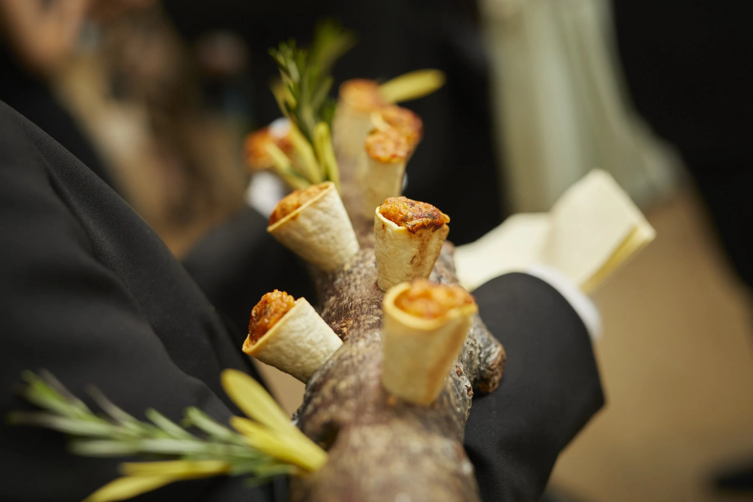 Close-up of a chef's hand holding an array of small cone-shaped appetizers topped with various ingredients, garnished with a sprig of rosemary, against a dark background.