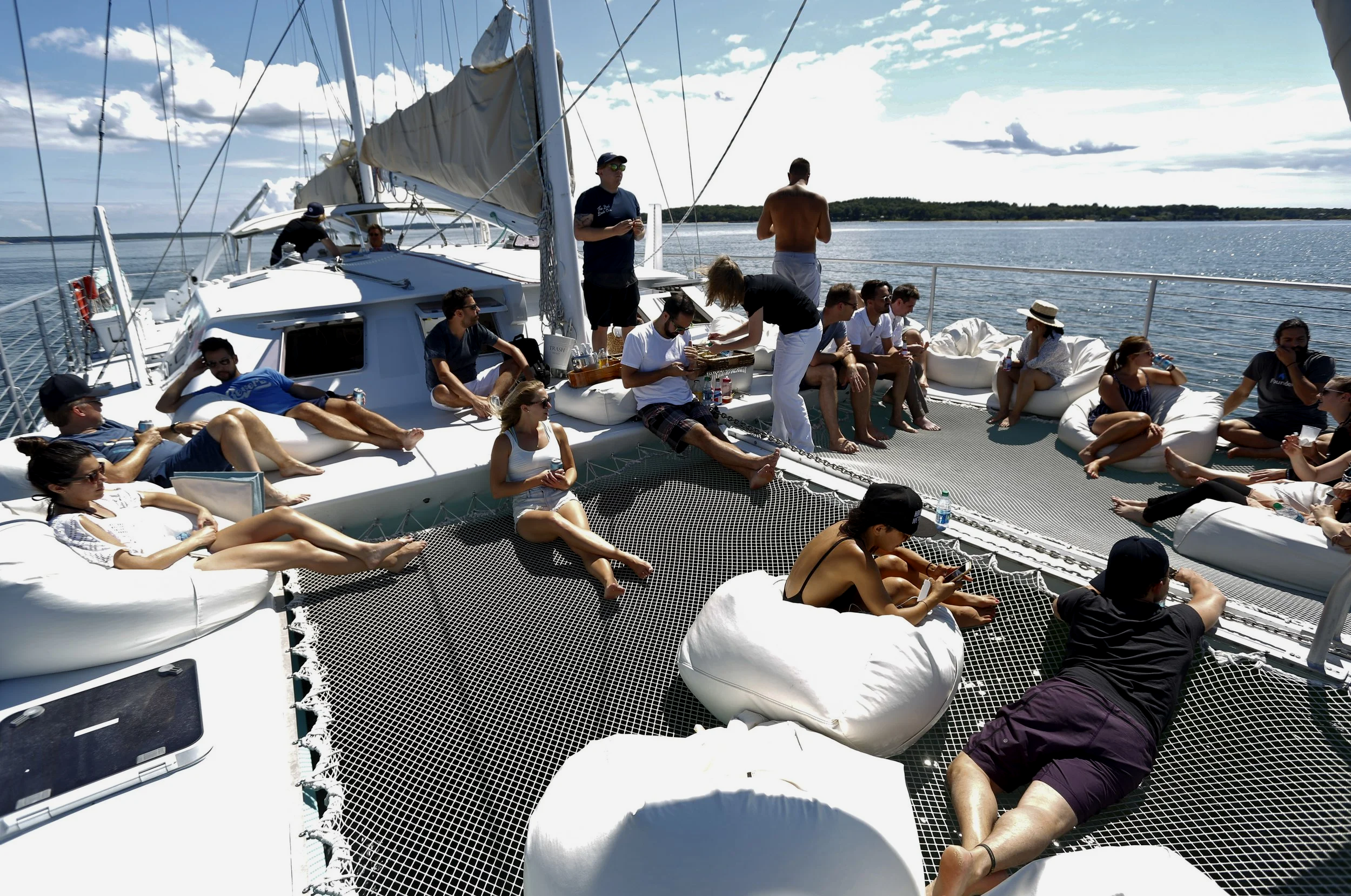 People relaxing on a boat deck with lounging cushions, some sitting and some lying down, enjoying a boat trip on the water on a sunny day with a cloudy sky.
