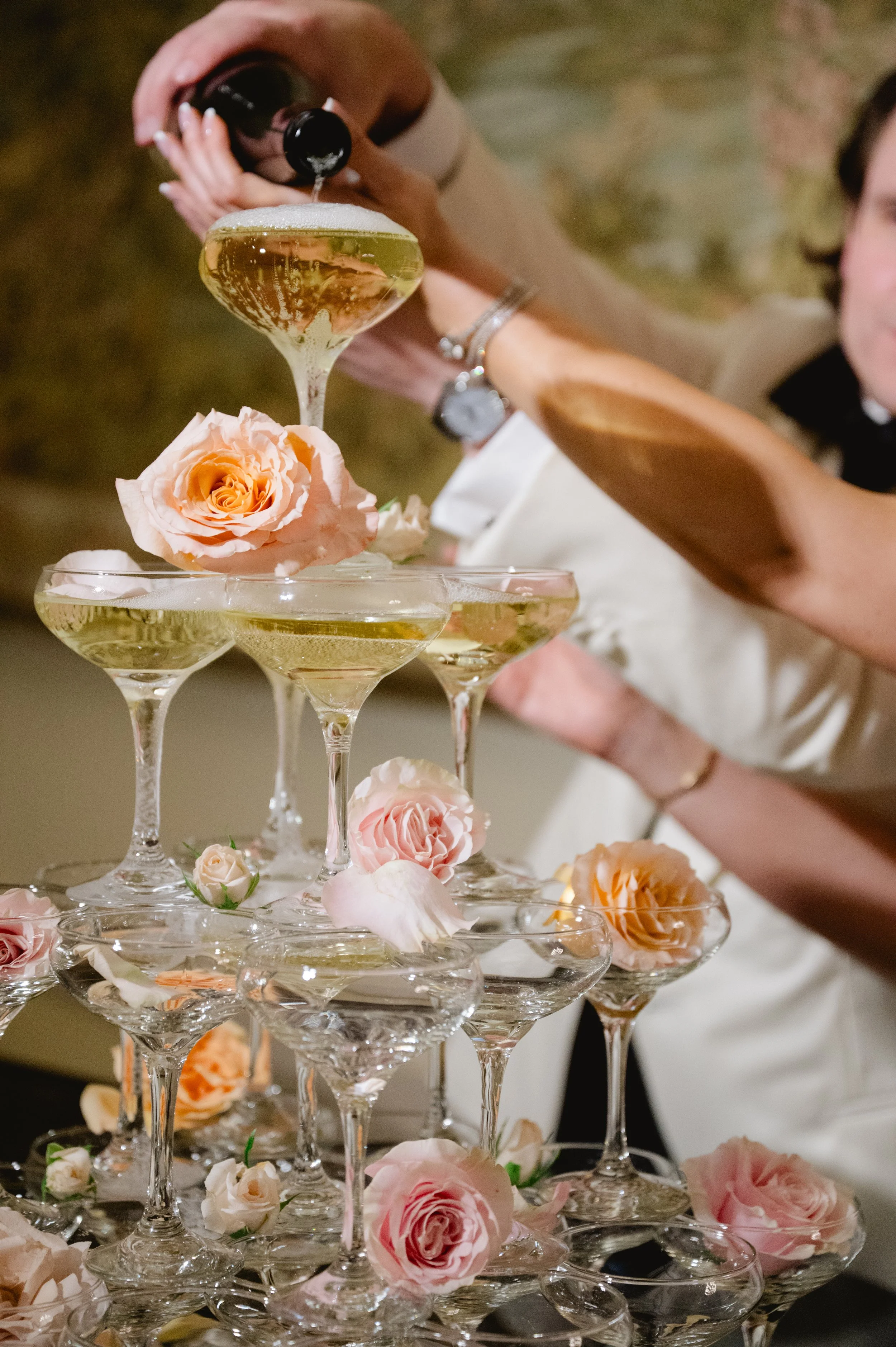 People pouring champagne into a tower of coupe glasses decorated with pink roses at a celebration or wedding.