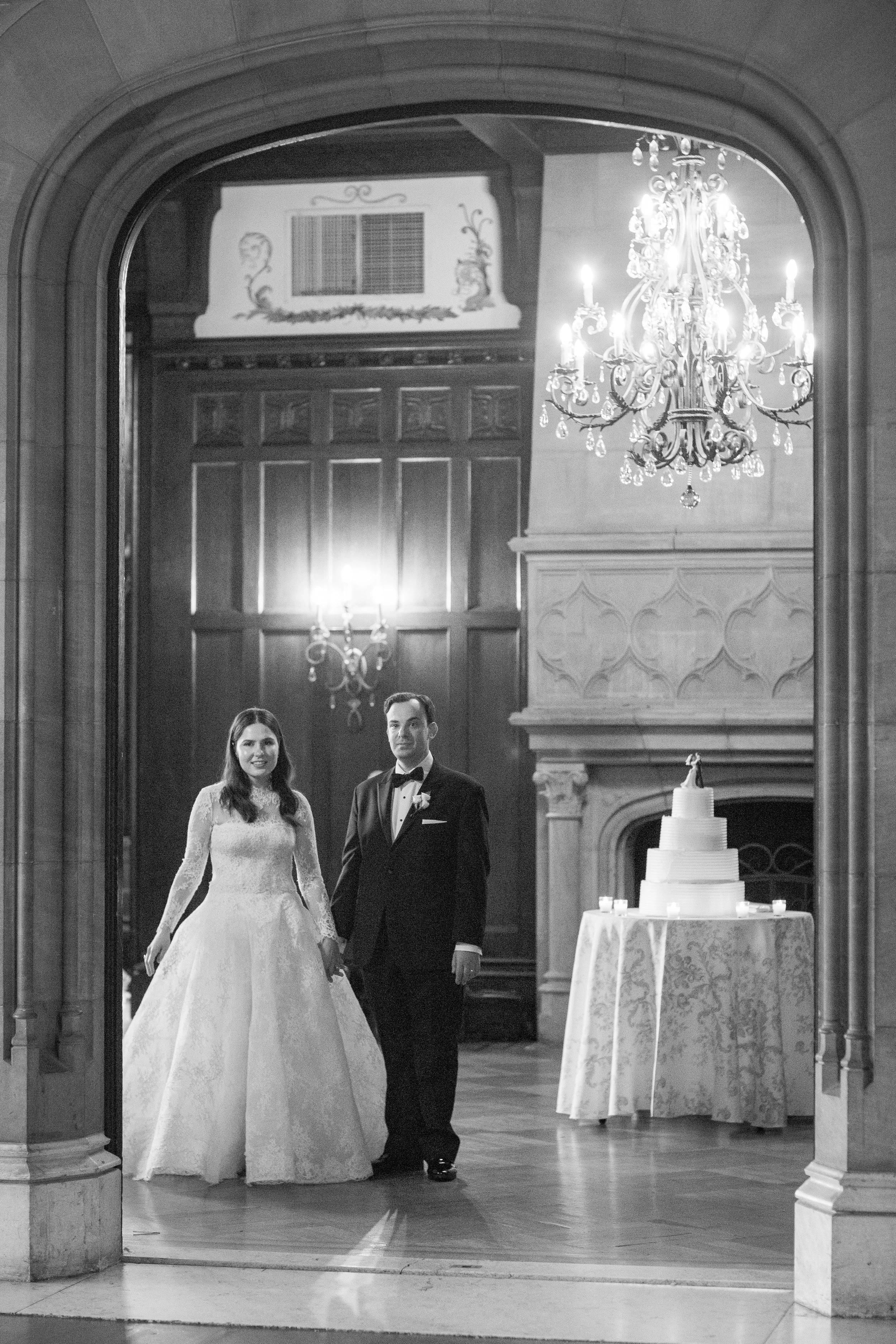 A bride and groom standing together at their wedding reception, smiling, with a wedding cake on a table to the right, chandelier overhead, in a grand hall with wooden paneling and decorative wall features.