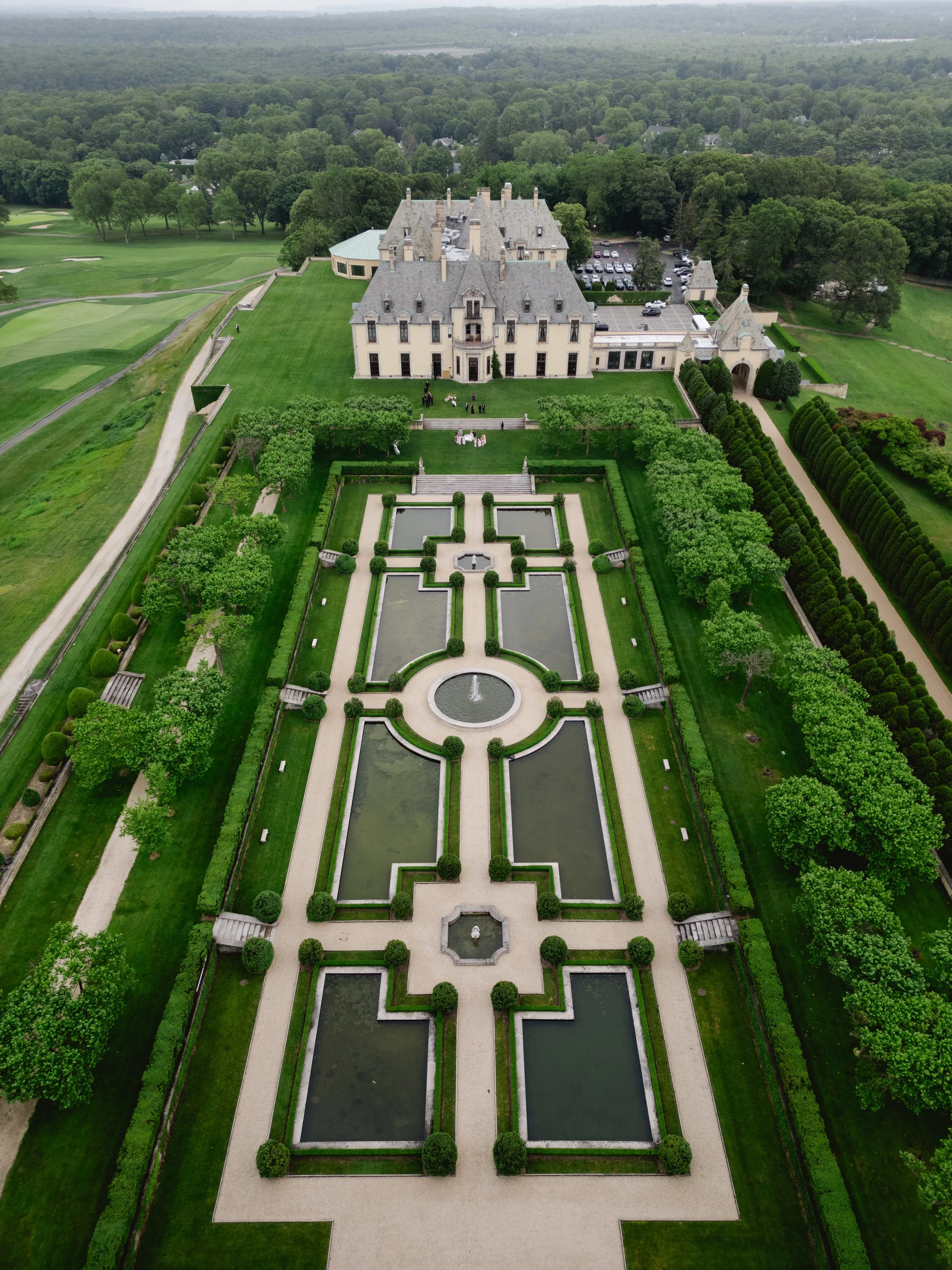 Aerial view of a large castle with a formal garden featuring symmetrical water features, pathways, and manicured bushes, surrounded by lush green lawns and trees.