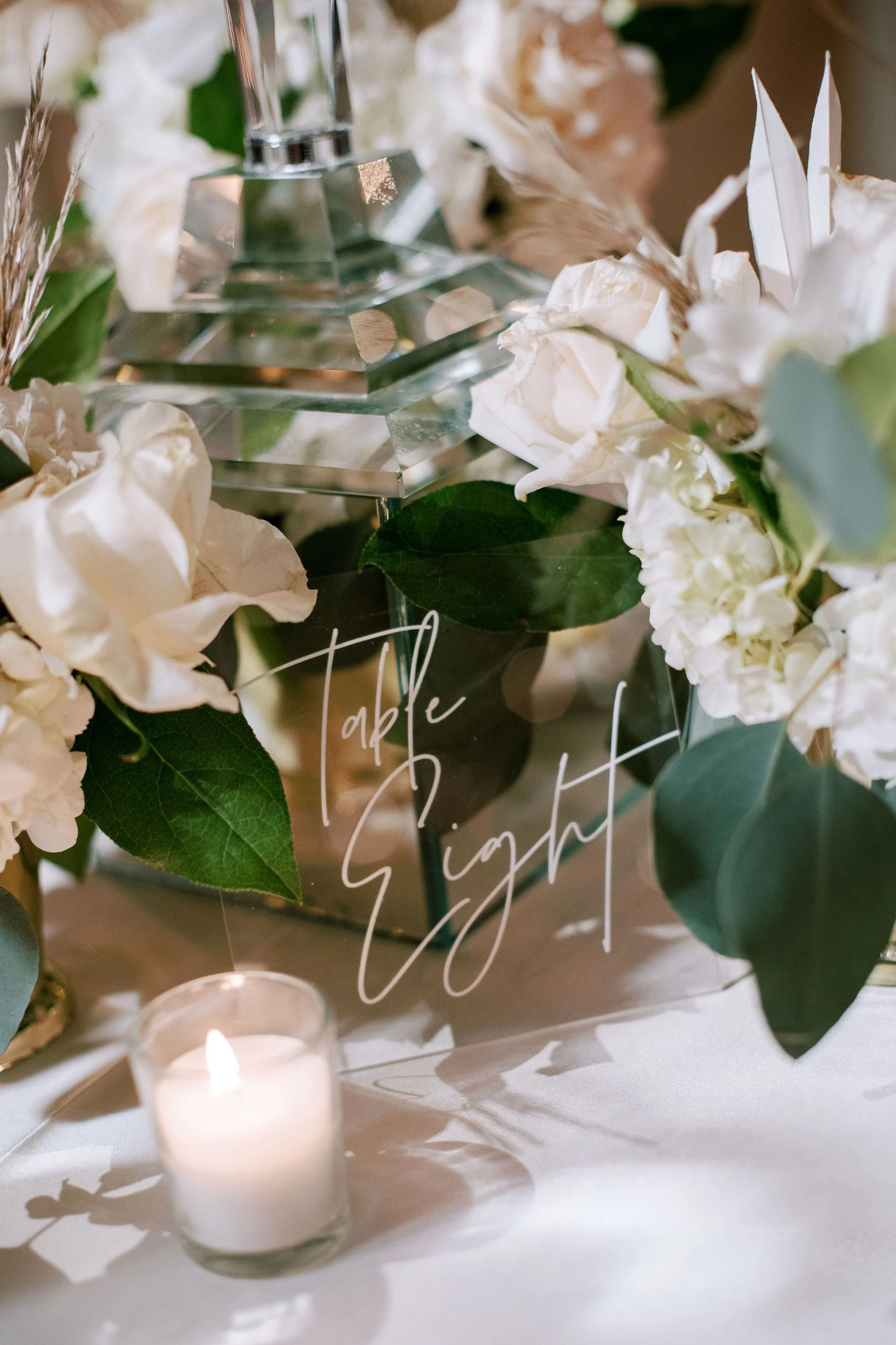 A close-up of a decorative arrangement with white flowers, green leaves, a lit white candle in a glass holder, a clear acrylic sign with white cursive writing, and a small candle in a glass holder on a white surface.