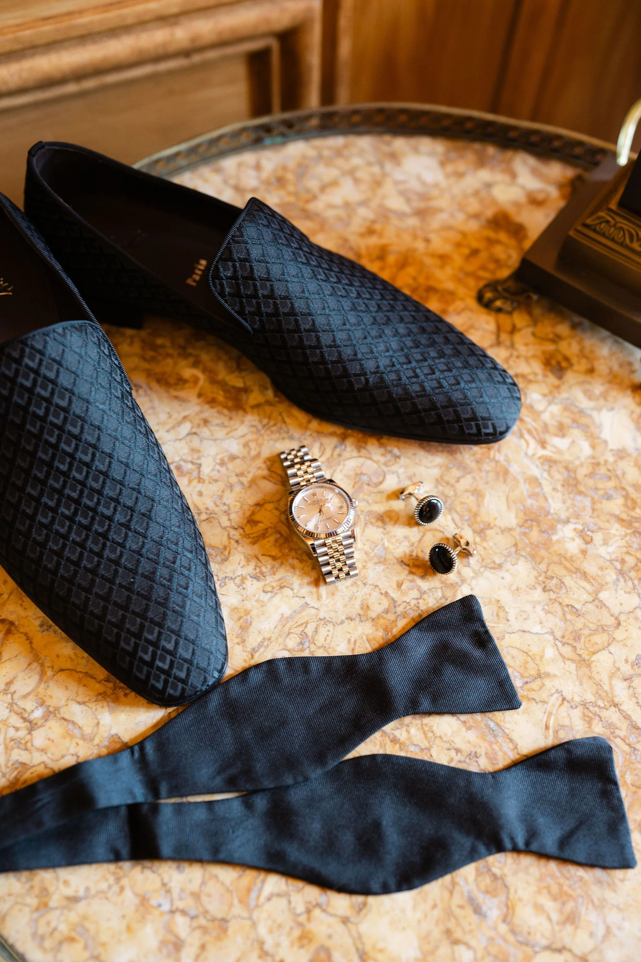 Black textured shoes placed on a marble surface, with a silver watch, a pair of black cufflinks, and a black ribbon nearby.