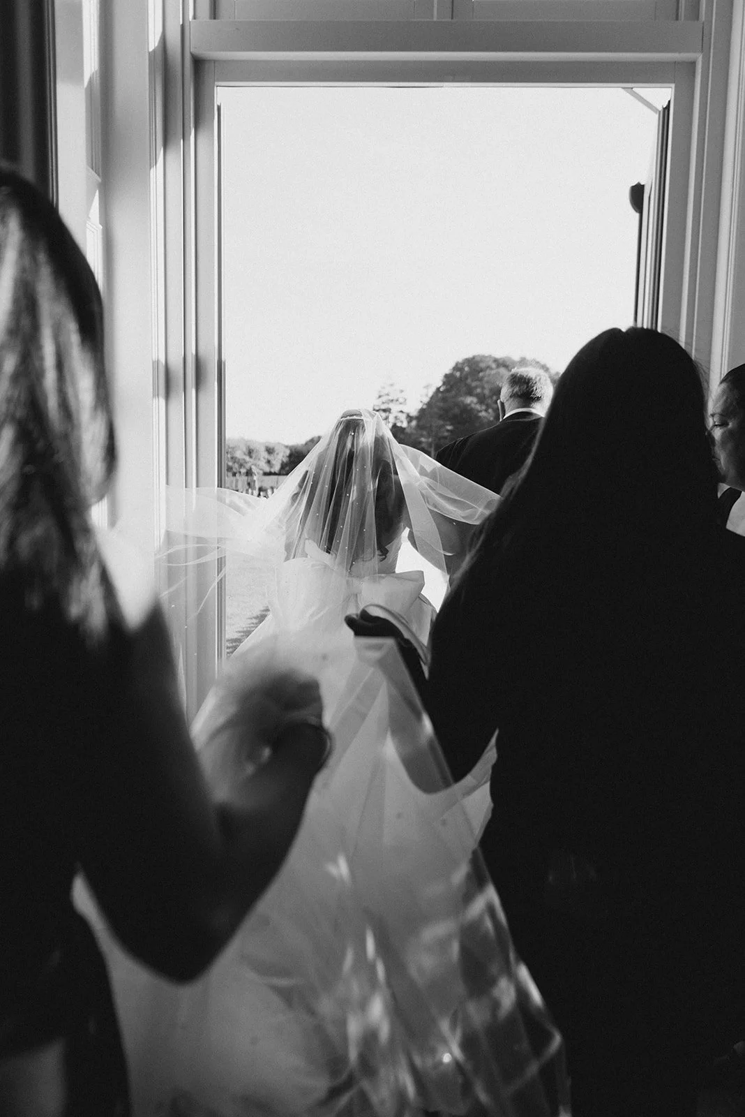 Black and white photo of a bride and groom walking out of a building during their wedding ceremony, seen through a doorway, with guests watching.
