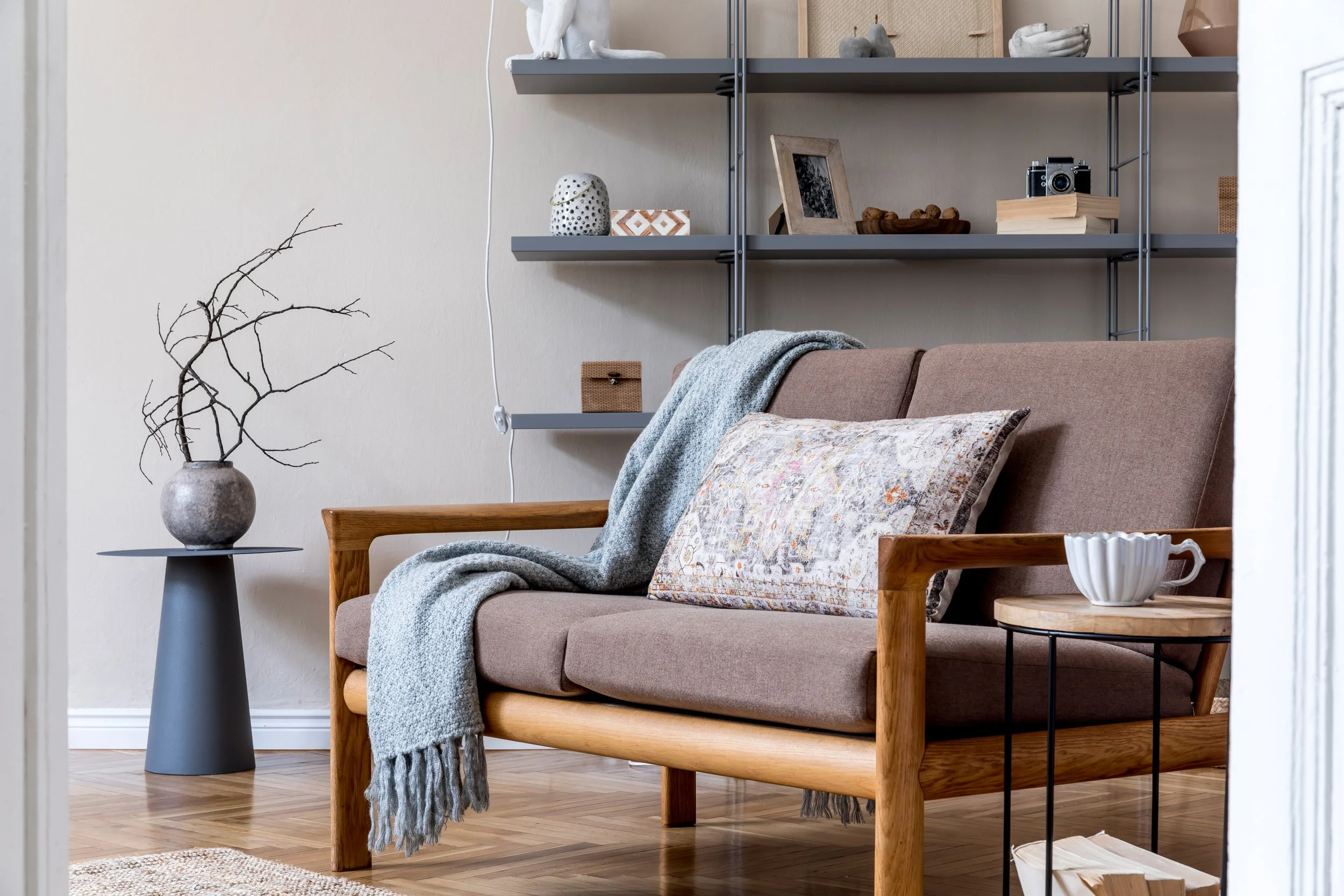 A cozy living room with a brown couch, a light gray blanket and a patterned cushion. Next to the couch is a black side table with a rustic vase holding leafless branches. Behind the couch, there is a gray shelving unit with decorative items, including a vintage camera, books, a framed photo, and small sculptures.