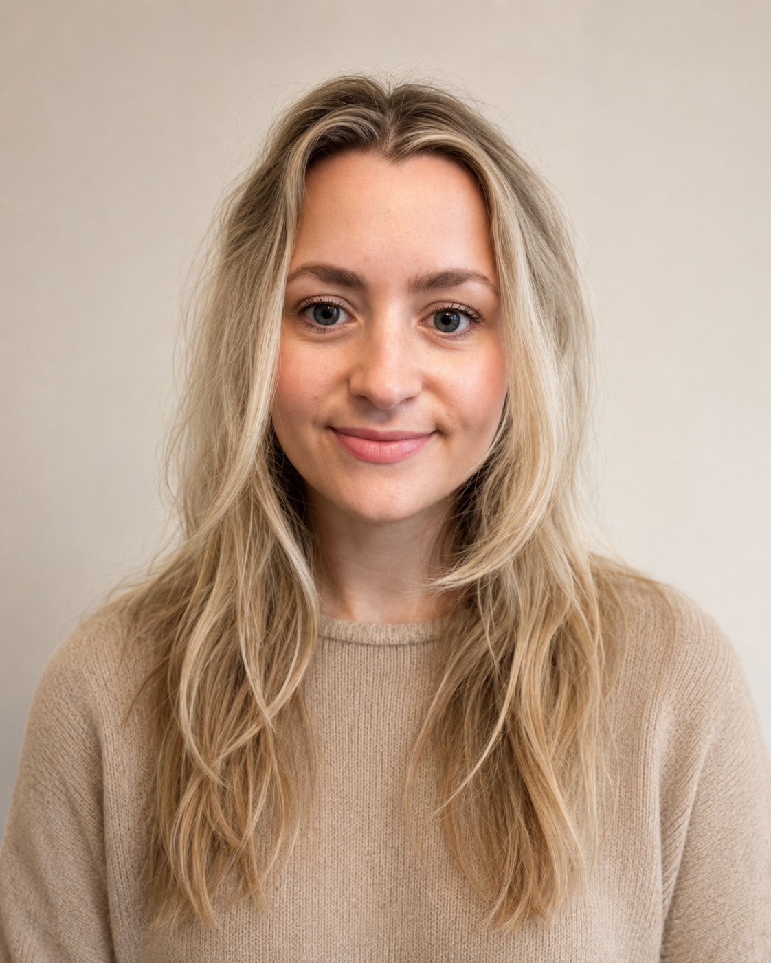 A young woman with long, wavy blonde hair and blue eyes, smiling gently at the camera, wearing a beige sweater, against a plain light background.