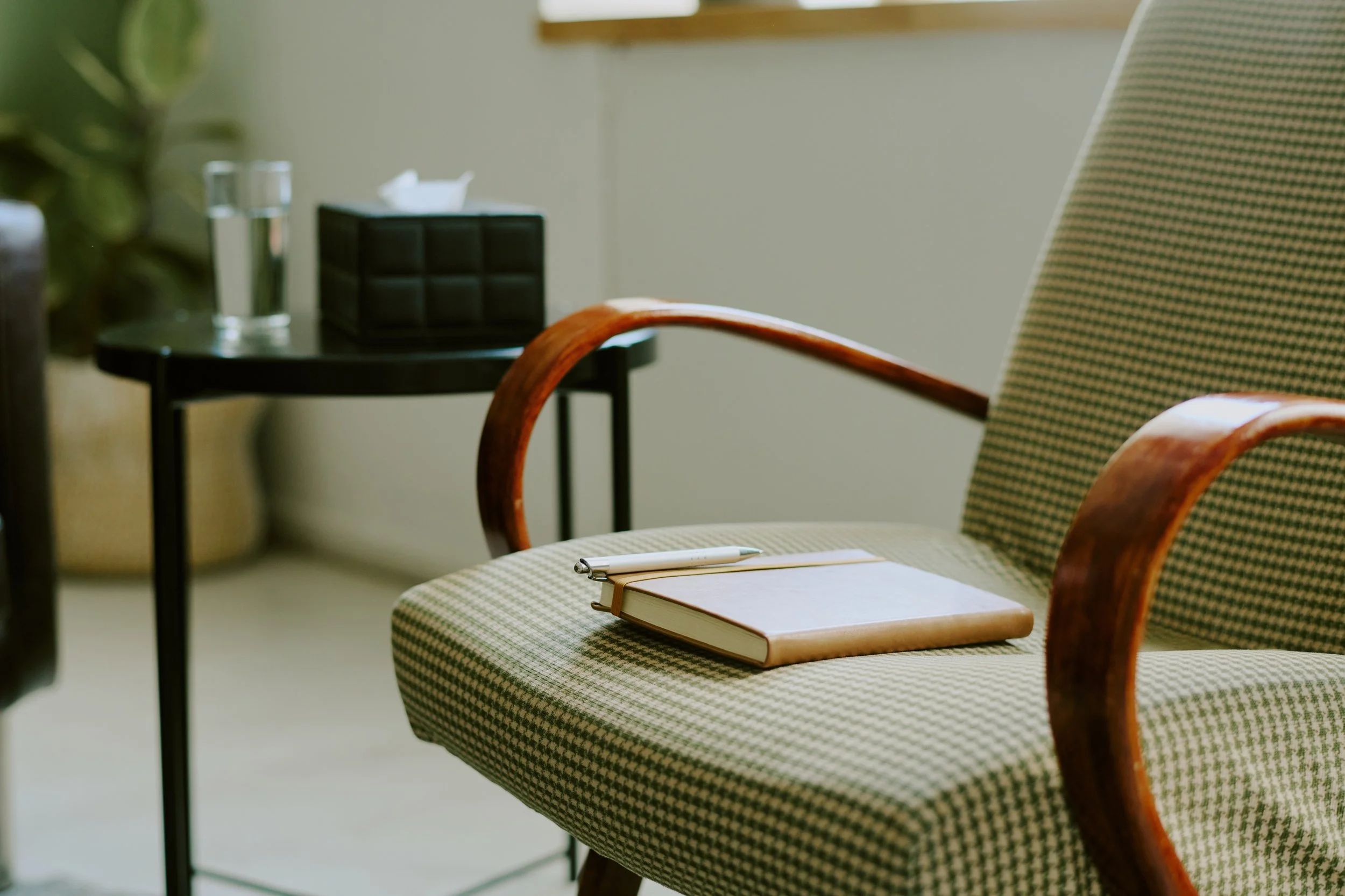 A cozy office space with a patterned armchair, a notepad and pen on the armchair, a small black side table, a glass of water, and a tissue box on the table.