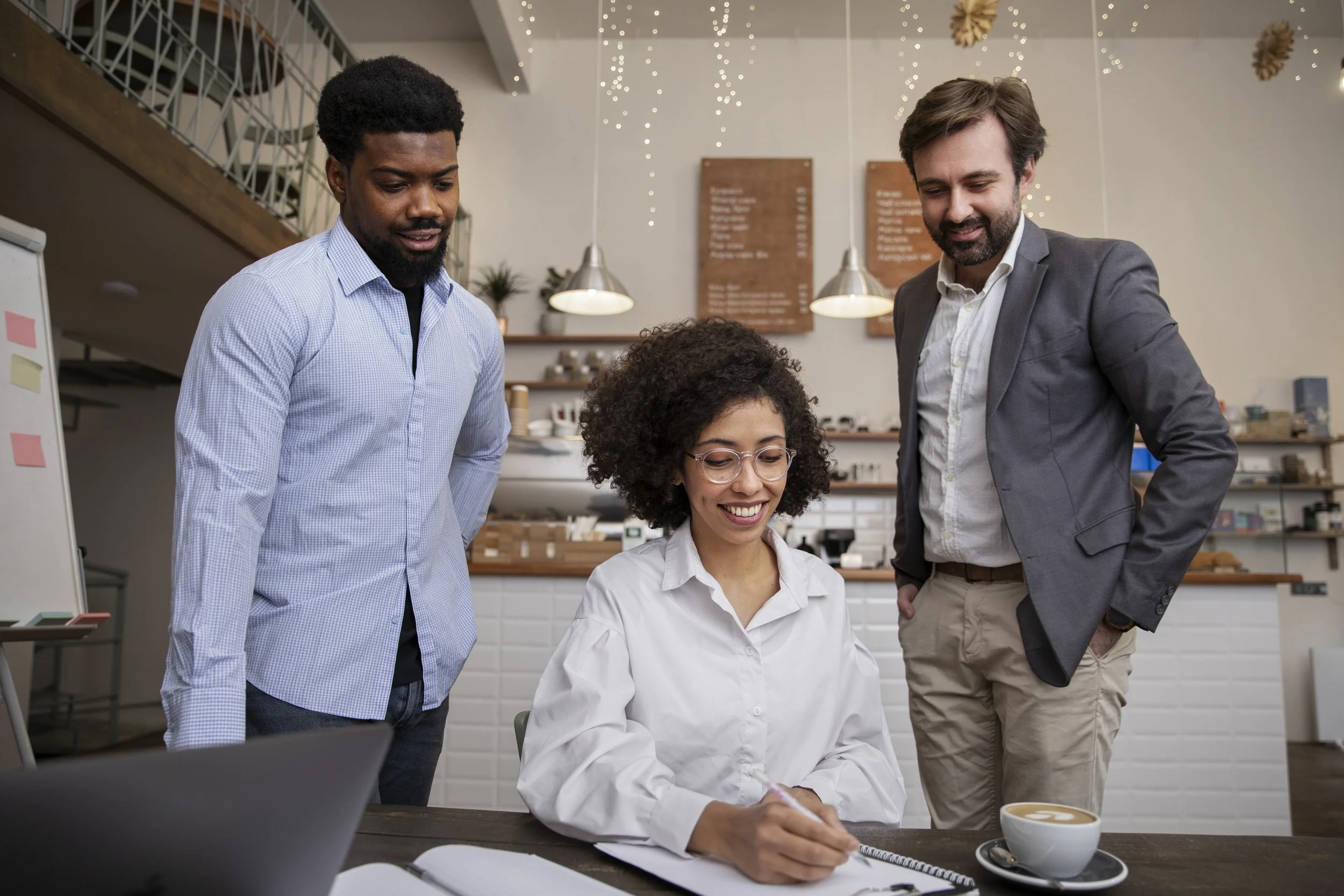 Quatre personnes dans un café ou un espace de bureau collaboratif. Deux hommes et une femme regardent un ordinateur portable, la femme sourit en écrivant dans un carnet. Un des hommes porte une veste grise, l'autre porte une chemise bleue. La scène est décorée avec des guirlandes lumineuses, des plantes, et des panneaux de menu en arrière-plan.