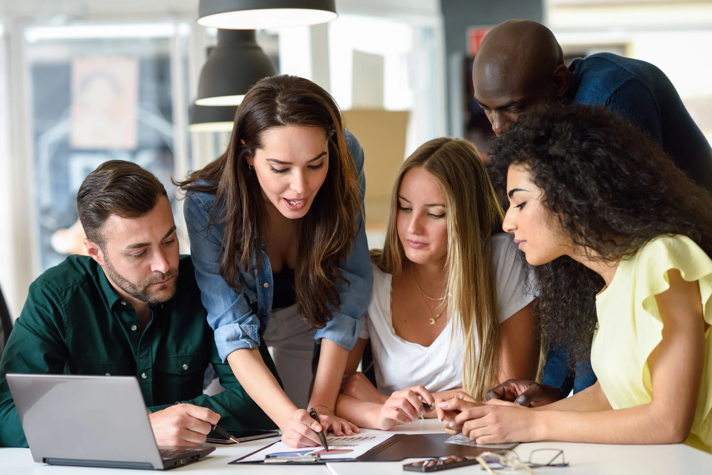 Groupe de jeunes adultes travaillant ensemble autour d'une table, collaborant sur un projet, avec des documents, un ordinateur portable et des stylos, dans un environnement lumineux.