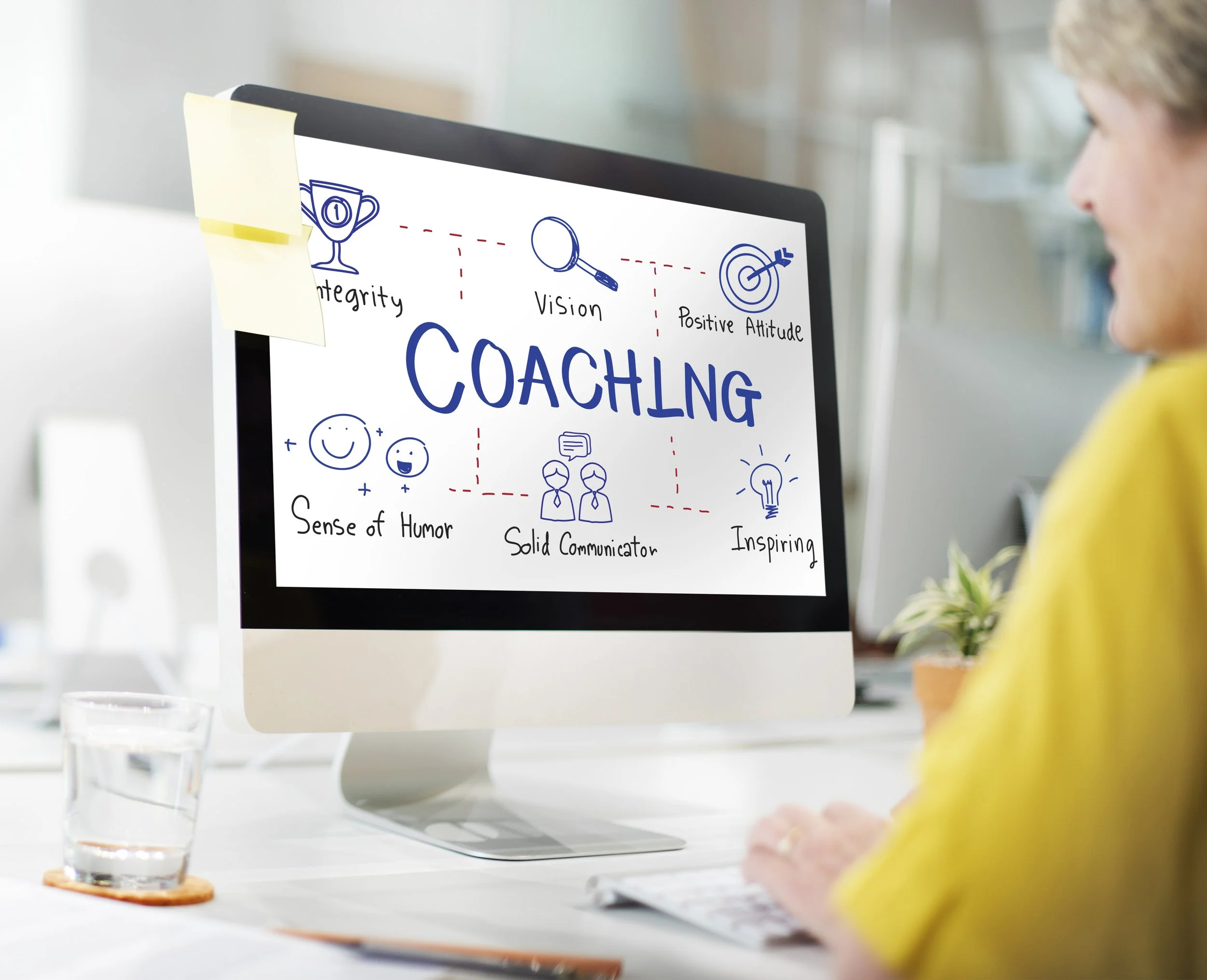 Un écran d'ordinateur affiche un tableau blanc avec le mot 'coaching' écrit en grand, entouré de dessins et de mots liés au coaching comme 'intégrité', 'vision', 'attitude positive', 'sens de l'humour', 'communicateur solide', et 'inspiration'. Un post-it jaune est collé en haut à gauche de l'écran. Une personne portant un vêtement jaune est assise devant l'écran, visible partiellement.