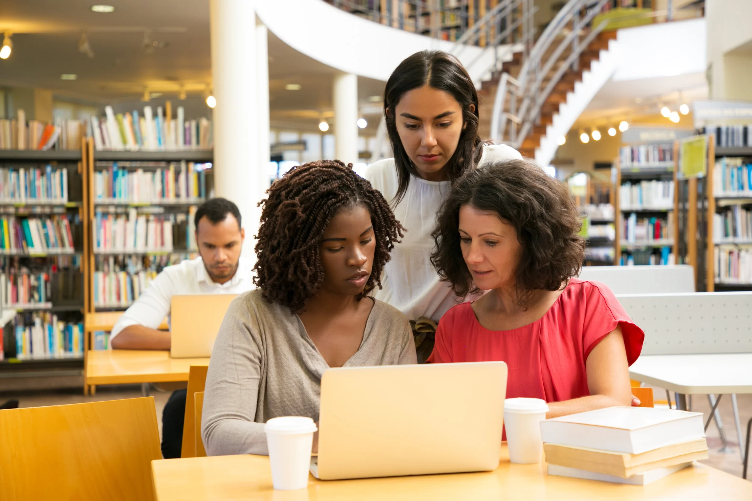 Groupe de cinq personnes dans une bibliothèque, deux femmes regardant un ordinateur portable, un homme concentré sur sa tâche, avec des étagères de livres en arrière-plan.