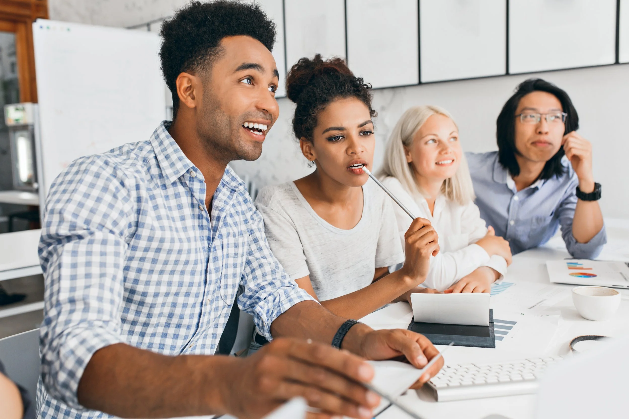 Groupe de quatre personnes assises à une table de réunion, dont un homme noir souriant, une femme noire utilisant un stylo, une femme blanche souriante, et une femme asiatique portant des lunettes. Ils semblent participer à une discussion ou une présentation dans un environnement de bureau moderne.
