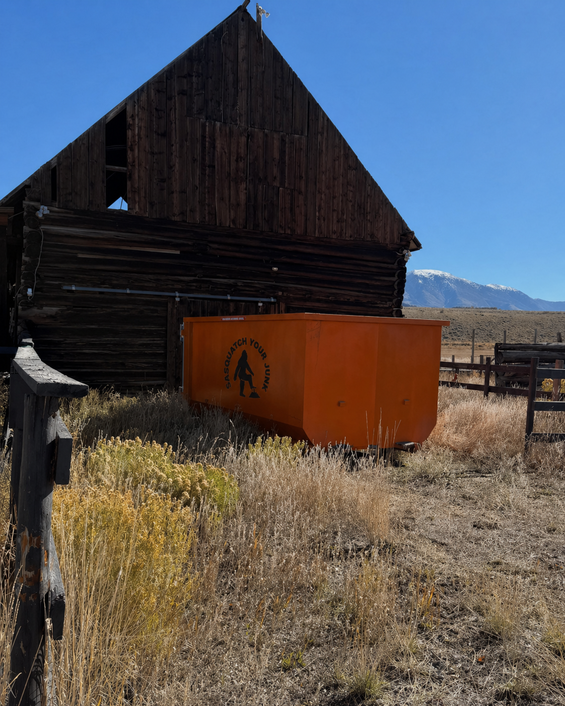 An old wooden barn with a weathered dark exterior, surrounded by dry grass, a bright orange container with a 'Sasquatch Your Junk' logo, and a distant snow-capped mountain under a clear blue sky.