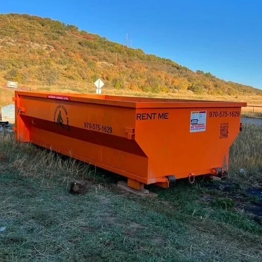 A large orange construction dumpster on a grassy field near a hill, with a park sign and trees in the background.