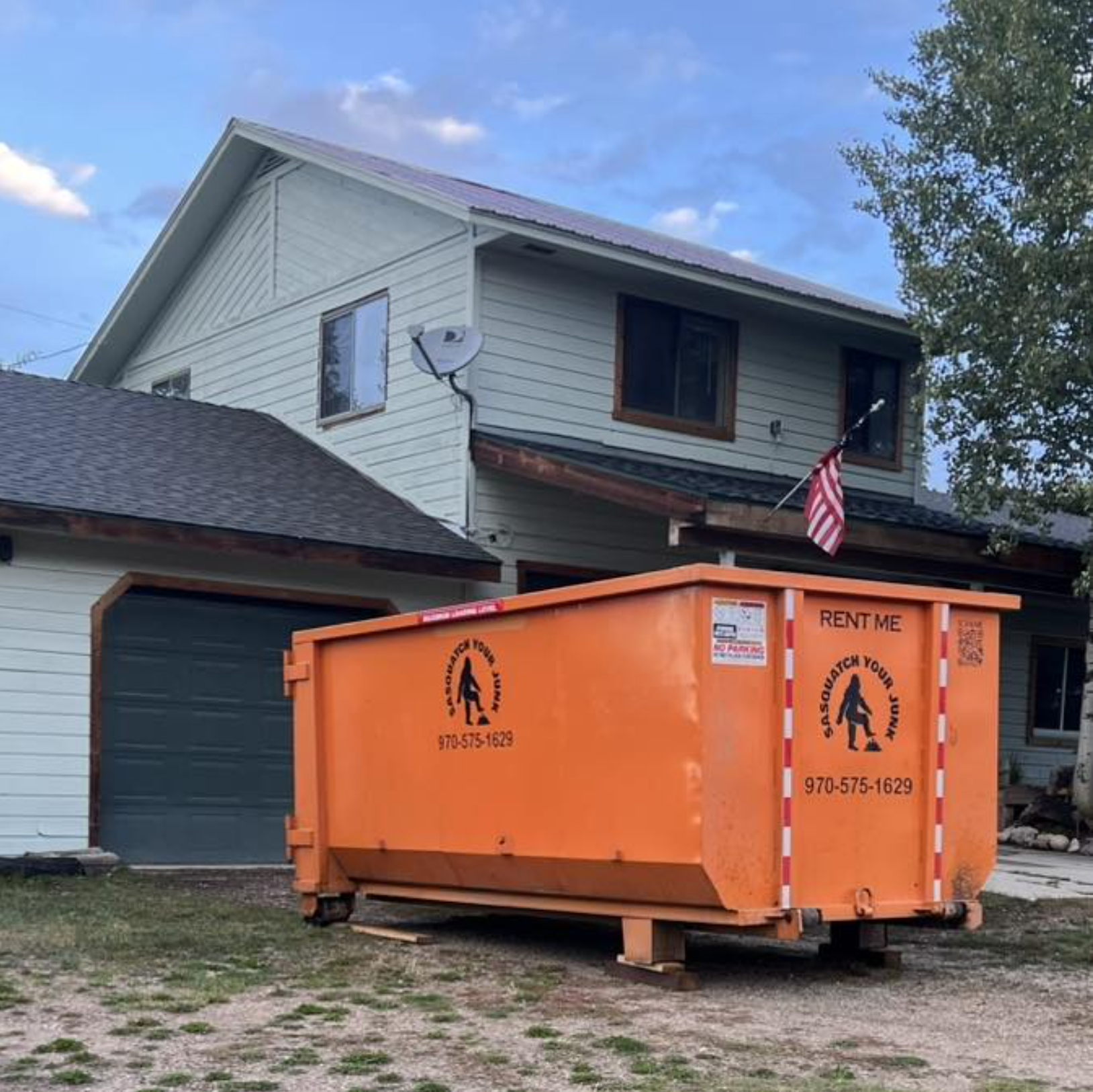 Orange trash dumpster in front of a house with two stories, beige siding, dark roof, and an American flag. House has window with wooden frame, satellite dish attached to the house, and a tree on the right side. The sky is partly cloudy.