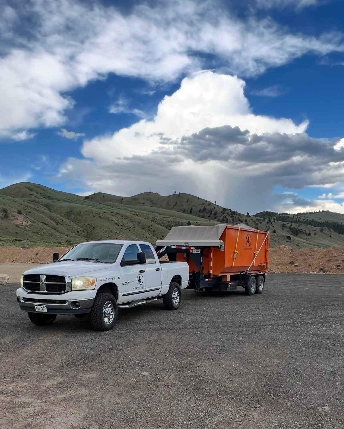 A white pickup truck with a logo on the door is parked on a dirt lot, attached to an orange trailer with a tarp cover. The background features green rolling hills and a sky filled with large, white and gray clouds.
