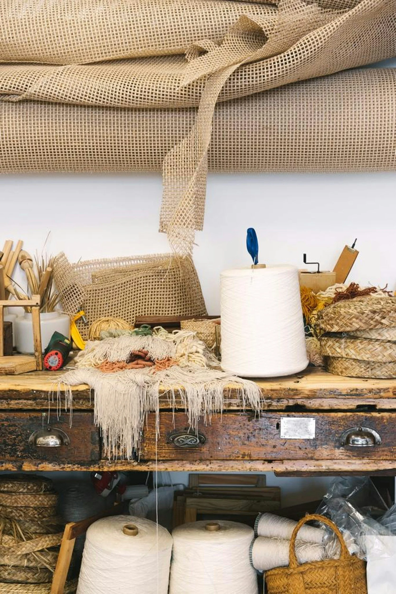 A cluttered workbench with various textiles, yarn, and weaving supplies, including spools of thread, woven baskets, and fabric.