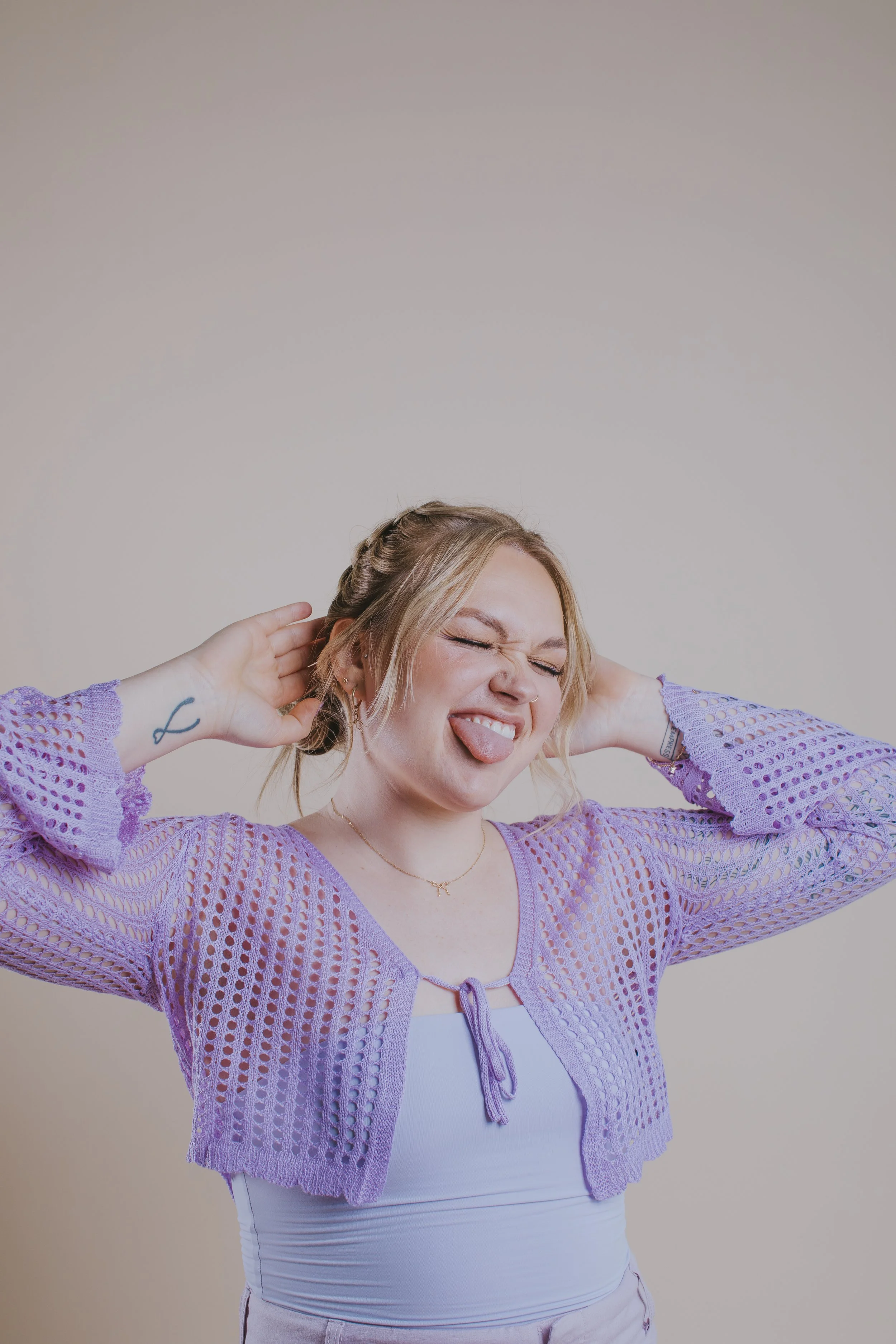 A young woman with blonde hair styled in braids, making a funny face with her eyes closed and tongue out, wearing a lavender crochet jacket over a white top and beige shorts, standing against a plain beige background.