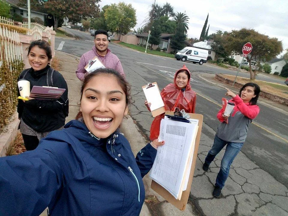 Group of five smiling people standing on sidewalk, holding clipboards, notebooks, and water bottles on an overcast day in a suburban neighborhood.