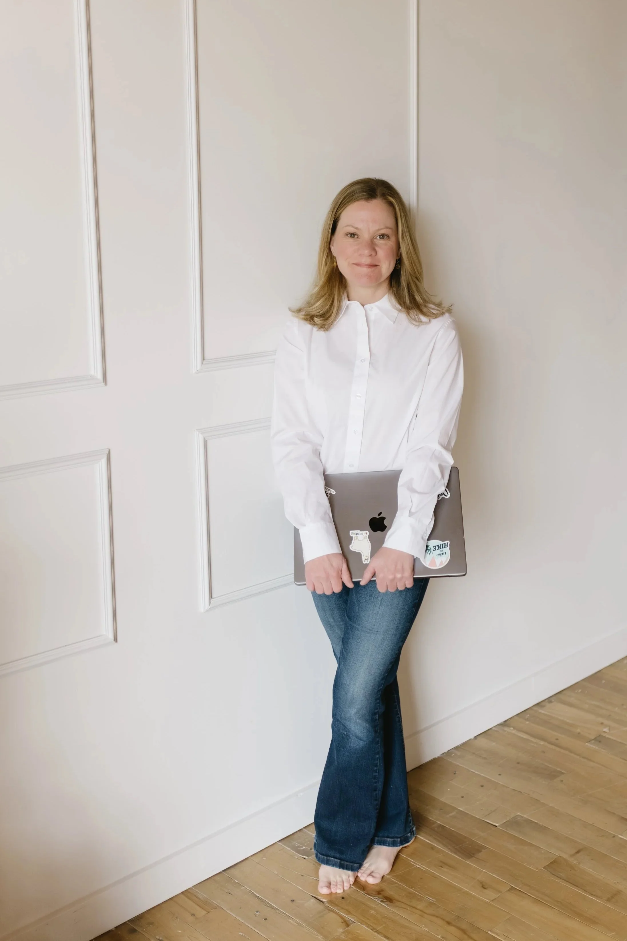 Woman standing barefoot indoors, holding a laptop covered with stickers, wearing a white blouse and blue jeans, leaning against a white wall with decorative panels, wooden floor.