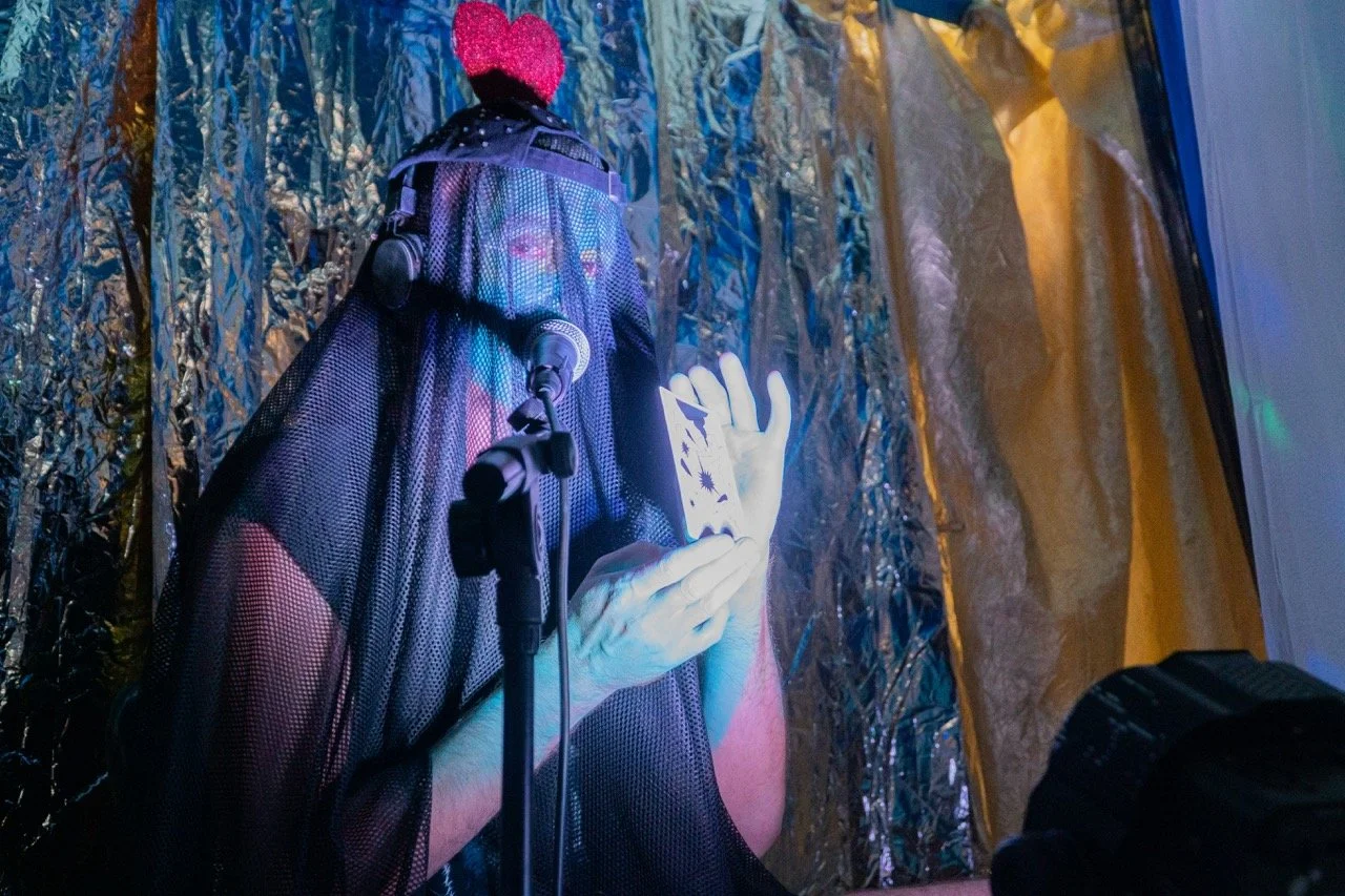 Une performance artistique où une personne est derrière un écran en tissu noir, portant un chapeau rose, et semble toucher ou manipuler un objet lumineux ou une fausse main. Le décor comprend des matériaux métalliques réfléchissants et une tente orange et blanche en arrière-plan.
