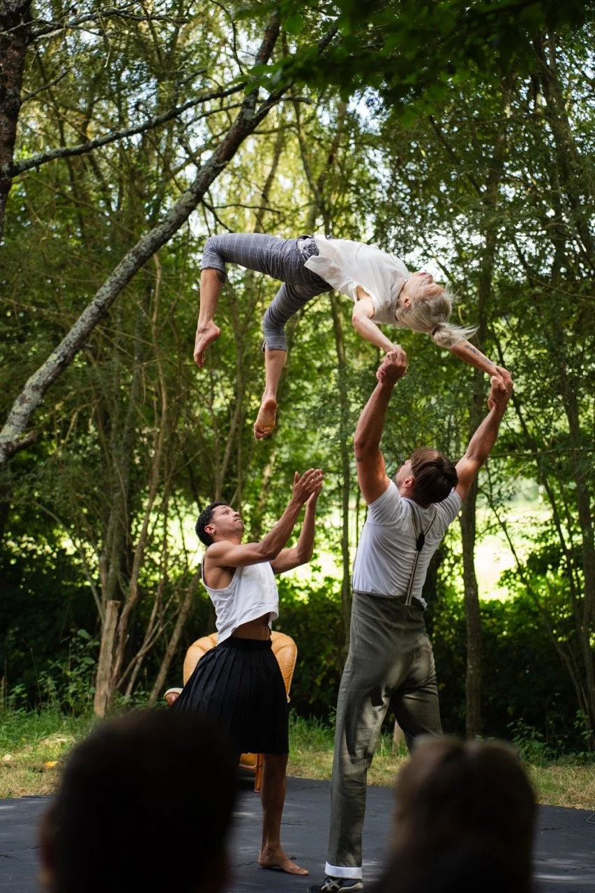 Deux hommes soulevant une femme dans les airs lors d'une performance de cirque ou de théâtre en plein air dans la forêt.