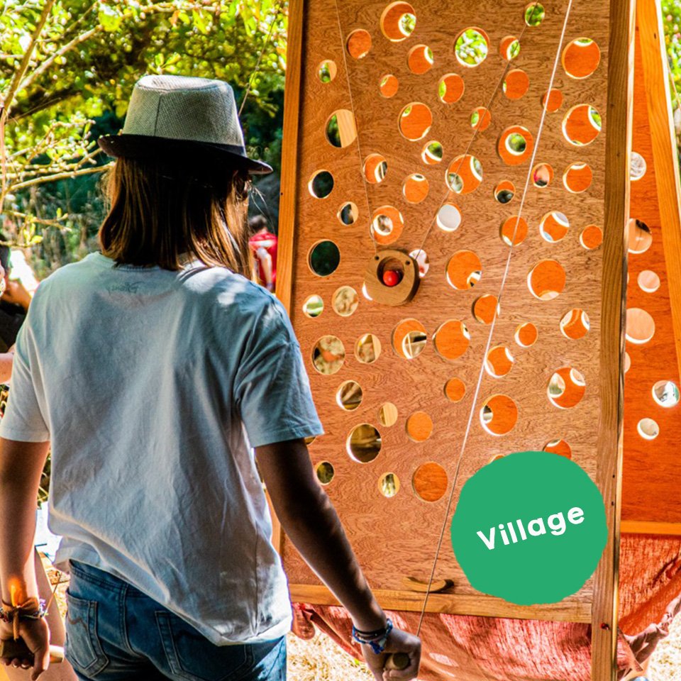 Une personne avec un chapeau regarde une grande planche en bois percée de nombreux trous, dans un environnement extérieur ensoleillé avec des arbres.