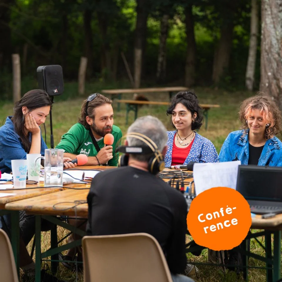 Une conférence en plein air avec cinq personnes assises autour d'une table en bois, en forêt. Deux femmes et un homme parlent dans un micro, tandis que deux autres femmes écoutent et sourient. Il y a des équipements audio et un ordinateur sur la table.