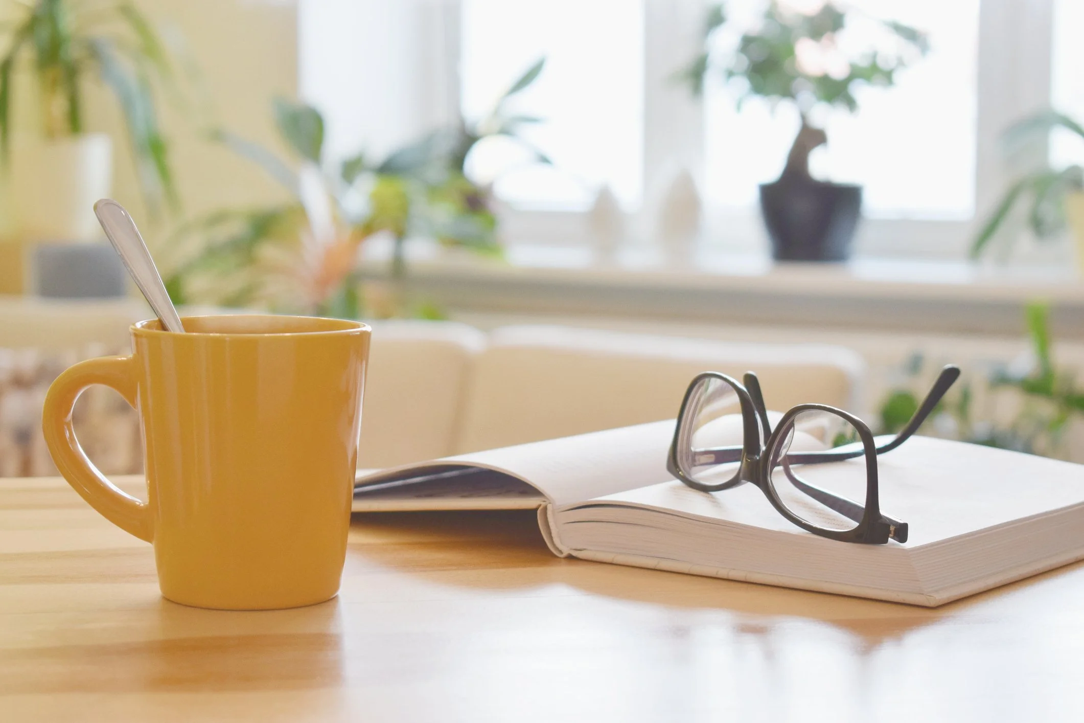 A yellow mug with a spoon inside, a pair of black-rimmed eyeglasses, a notebook or magazine, and a pen sit on a wooden table. In the background, a window with blurred green plants on the windowsill is visible.