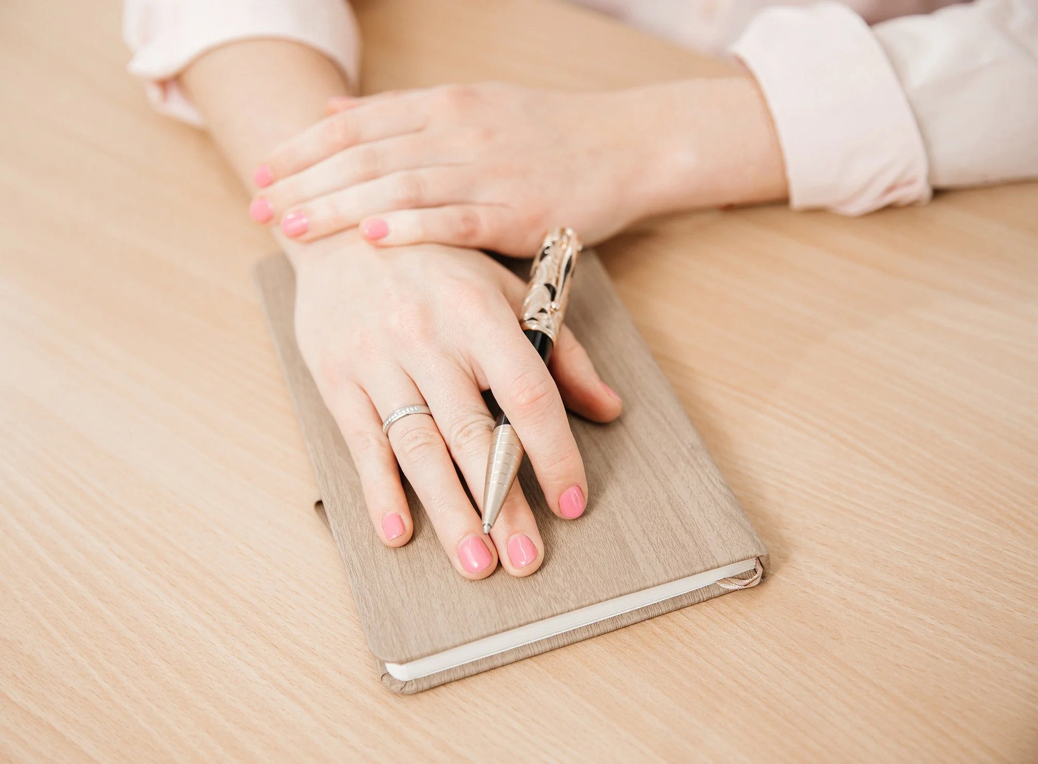 A person with pink painted nails wearing rings is writing in a notebook with a gold pen, resting their hand on top of another person's hand on a wooden desk while in EMDR individual consultation.