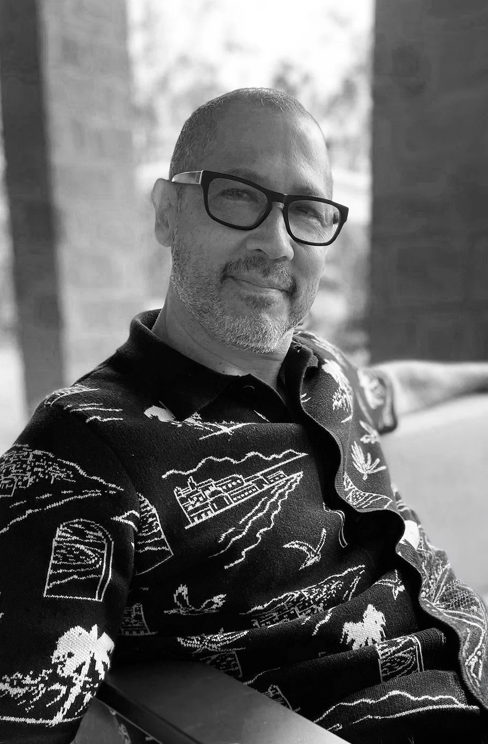 A man with glasses and a beard smiling, sitting on a porch or terrace in front of a window, wearing a patterned shirt with various outdoor scenes and animals.